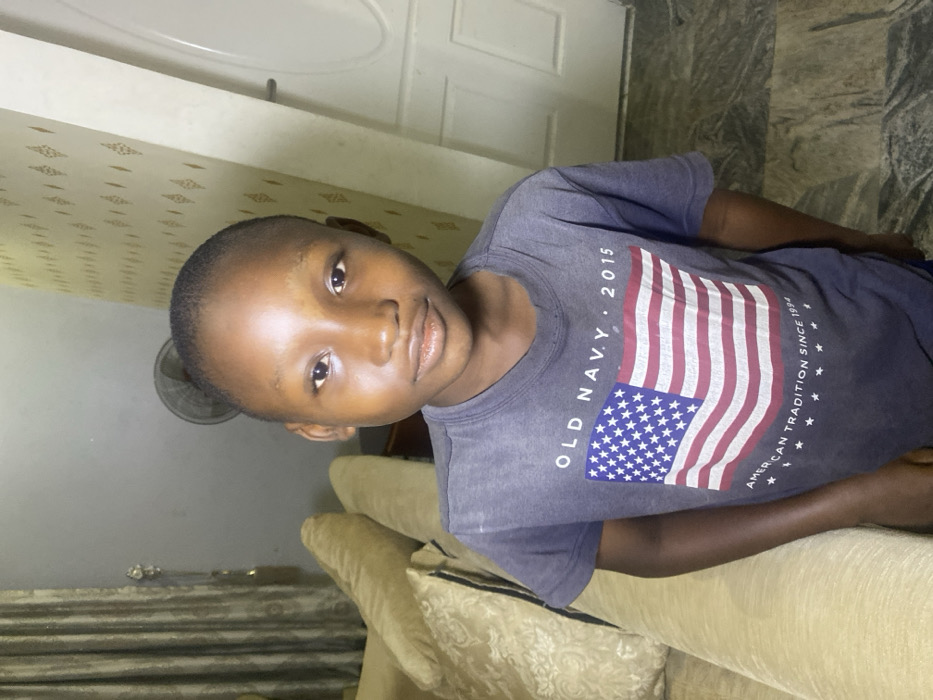 A young boy is resting on a beige sofa. He has dark skin, short dark hair and is wearing a gray t-shirt with an American flag design. The shirt says "Old Navy 2015 American Tradition Since 1994". Behind him are a white door and patterned wallpaper. A fan is partially visible. The lighting is dim.