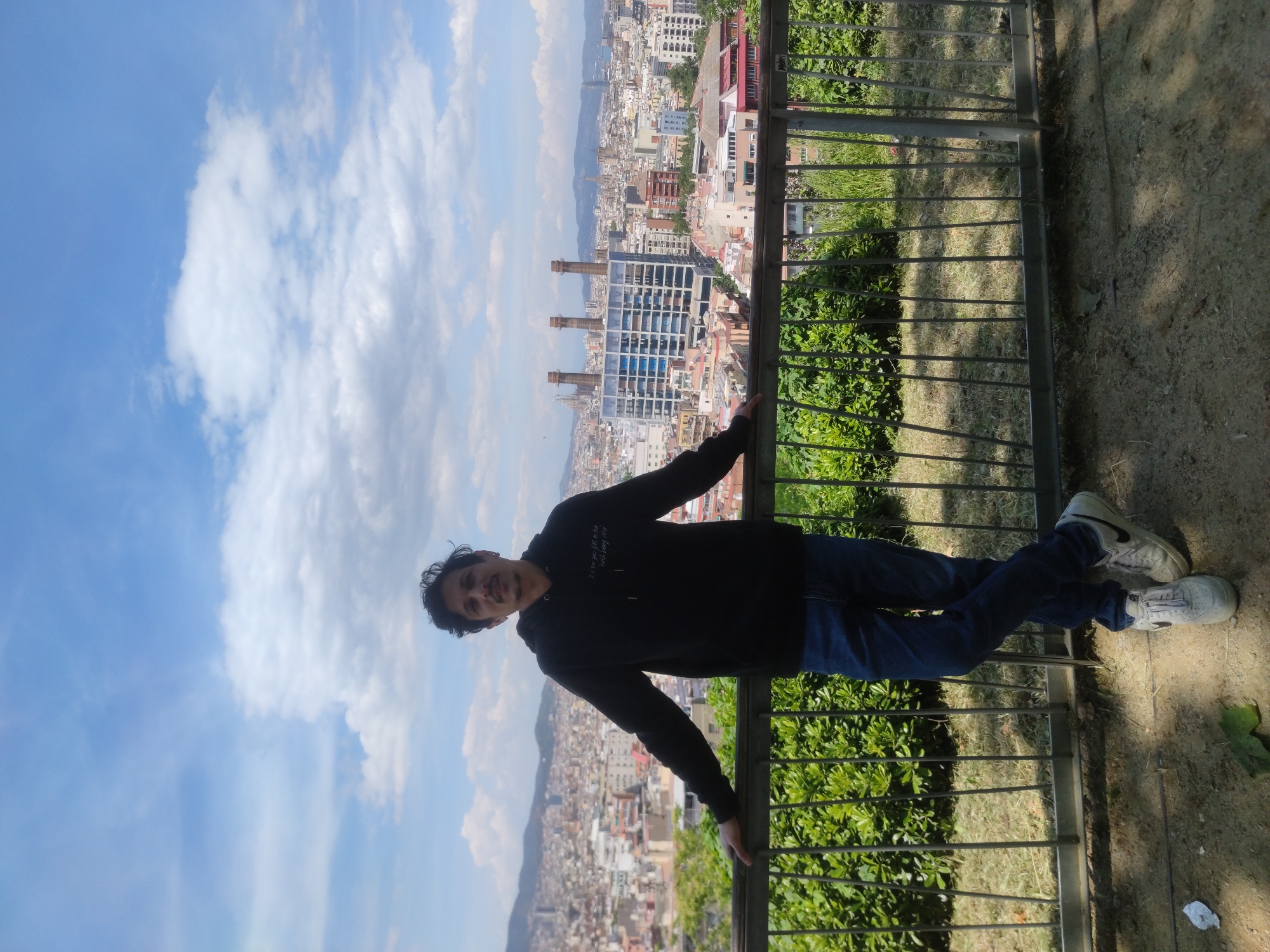 A man in a dark hoodie and jeans stands at a railing overlooking Barcelona, Spain. The city extends into the distance, with the blue sky and white clouds visible in the background. The railing is gray metal with green vegetation visible behind it.