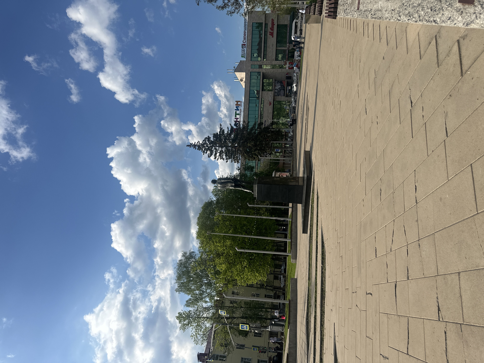 The image depicts a public space in the city of Naro-Fominsk, Russia. The scene features a wide pedestrian walkway paved with rectangular blocks. A statue on a plinth is partially visible amidst trees and lamp posts. A multi-story building with signs in Russian can be seen in the background; one sign includes the words "Супермаркет" (supermarket) and the brand "Мираторг". A blue sky with white clouds is overhead. In the lower part of the frame, there are trees, buildings and traffic signs. There is no activity occurring in the image.
