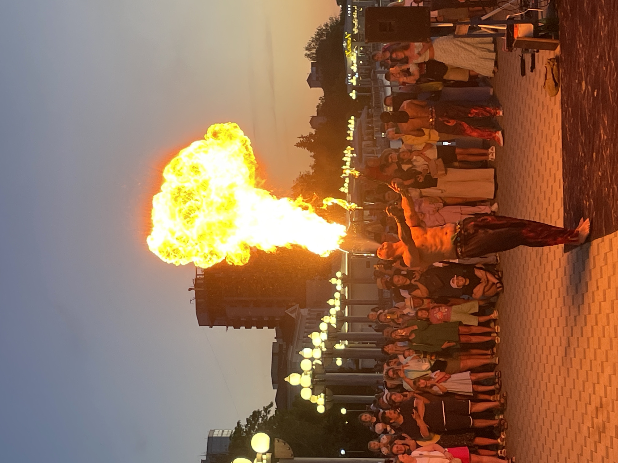 A male fire performer is the central subject, exhaling a large plume of fire and smoke upwards, illuminated by the setting sun. He is shirtless, wearing patterned pants, and stands on a paved area in front of an audience. The audience, composed of men, women, and children, watches the performance, many holding up phones to record. Behind the audience, a row of ornate lampposts with yellow lights lines a walkway. In the background, several buildings of varying heights and architectural styles are visible, with trees and hills beyond. The sky is a gradient of twilight colors, transitioning from orange near the horizon to blue. The scene appears to be set in a public outdoor space, possibly a promenade or park, in Anapa, Russia.