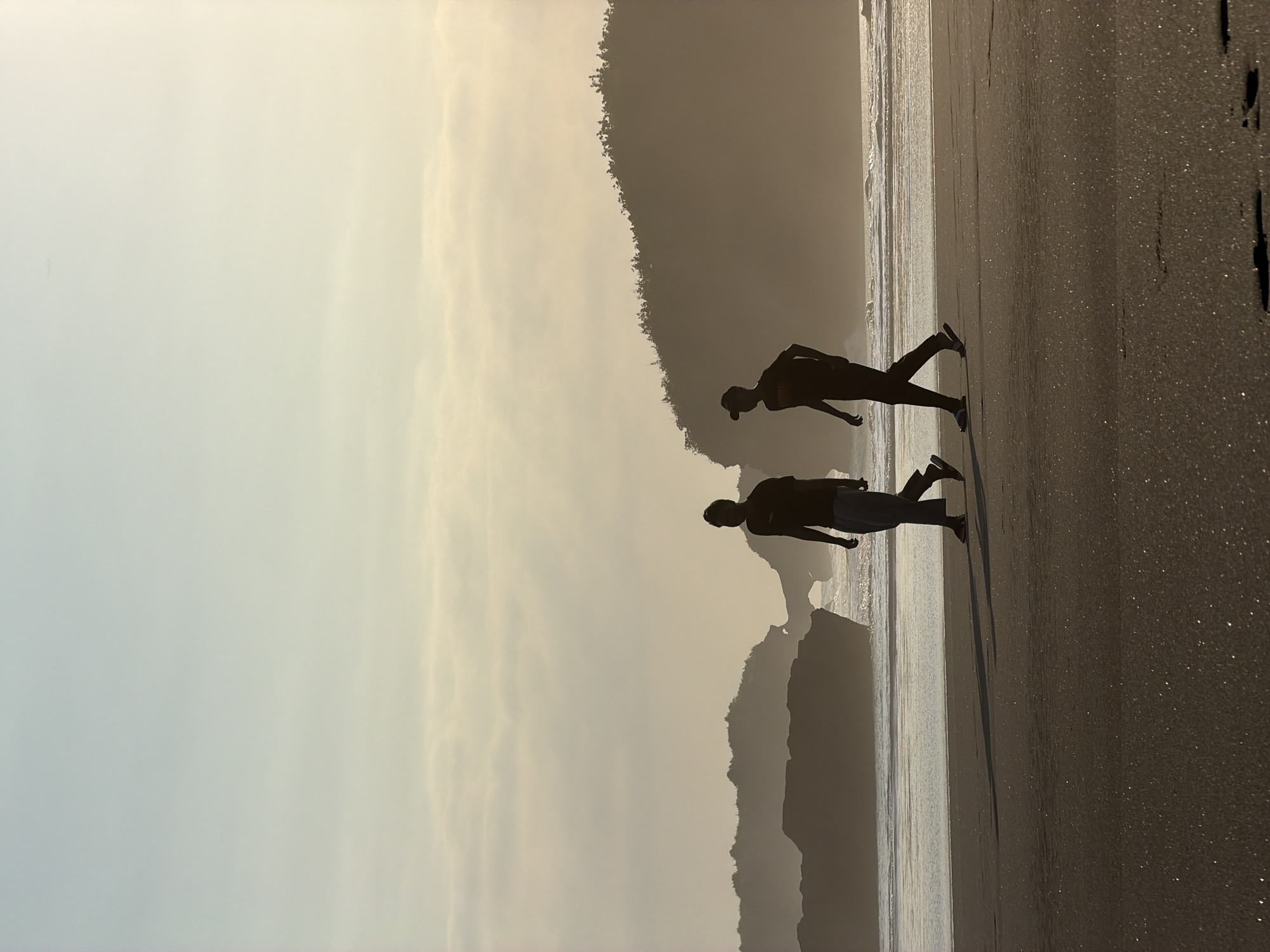 The image displays a coastal scene at a beach in Selemadeg, Indonesia, bathed in the soft, warm glow of either sunrise or sunset, which silhouettes the foreground subjects and distant landforms. Two individuals are walking from left to right across a wide expanse of dark, damp sand in the midground. The person on the left is taller, attired in a t-shirt and loose-fitting pants, and appears to be barefoot or wearing open-toed footwear. The person on the right, slightly shorter, wears a t-shirt, pants, and a baseball cap, with sandals or slippers on their feet. Both individuals cast elongated shadows forward onto the glittering sand.
In the background, the ocean features gentle waves breaking into white foam along the shoreline. Beyond the water, the horizon reveals distinct geological features: to the left, multiple dark, silhouetted rock formations or islands are visible, including a prominent natural rock arch. To the right, a large, dark, undulating hill or coastal mountain, densely covered in vegetation, rises from the ocean's edge. The sky transitions from a light blue at the top to a pale golden yellow near the horizon, with subtle, horizontally stretched cloud formations visible.