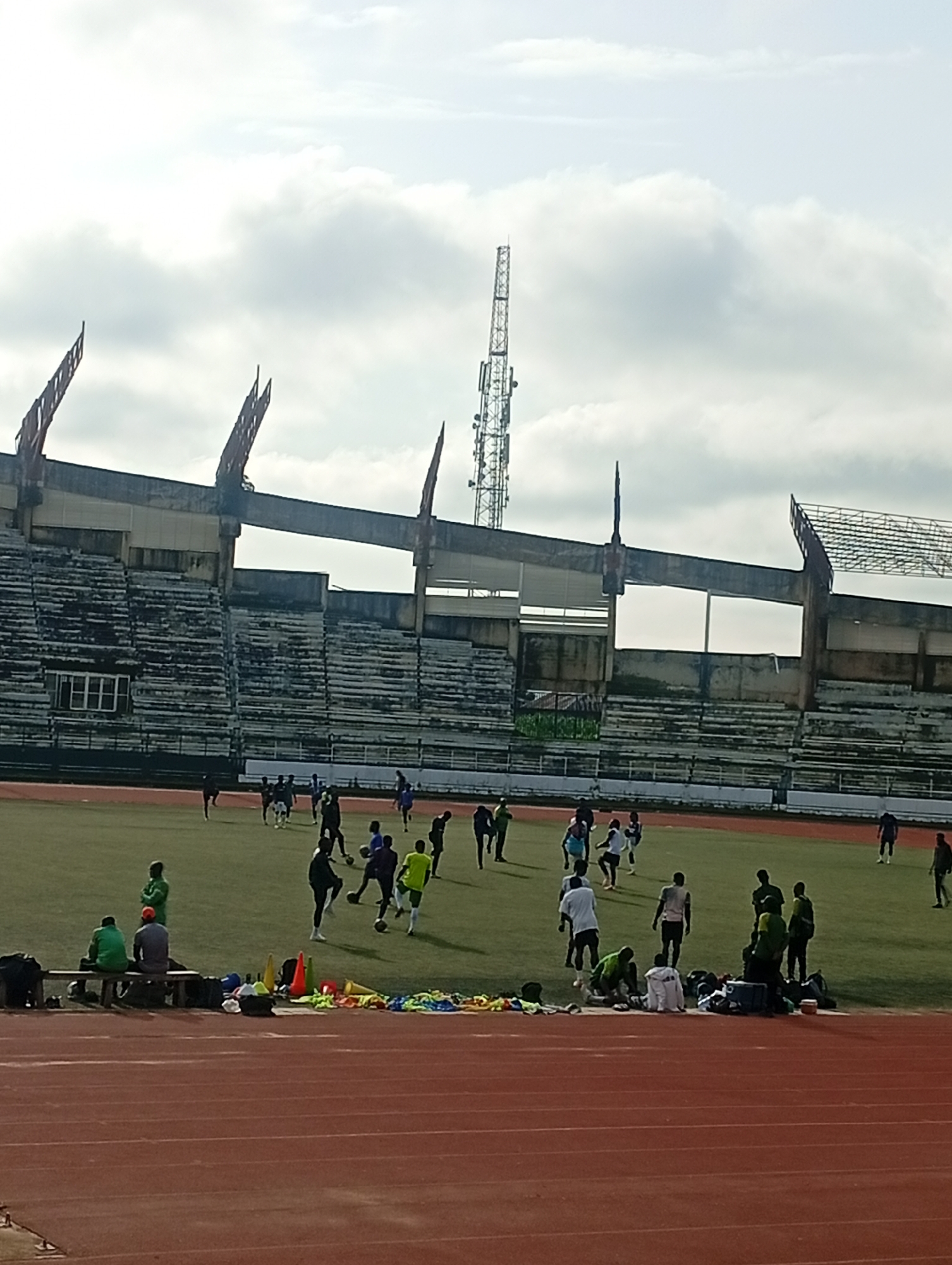 A wide shot captures an outdoor sports stadium with a soccer field. The field is green, and a track runs in the foreground. Several individuals, presumably soccer players, are scattered across the field, some of them actively engaged in a soccer training session, while others appear to be observing from the sidelines. The stadium seating is visible in the background, appearing weathered and partially occupied by a few spectators. The sky above is overcast with scattered clouds. The overall scene depicts a typical day at a soccer stadium during a practice or training session.