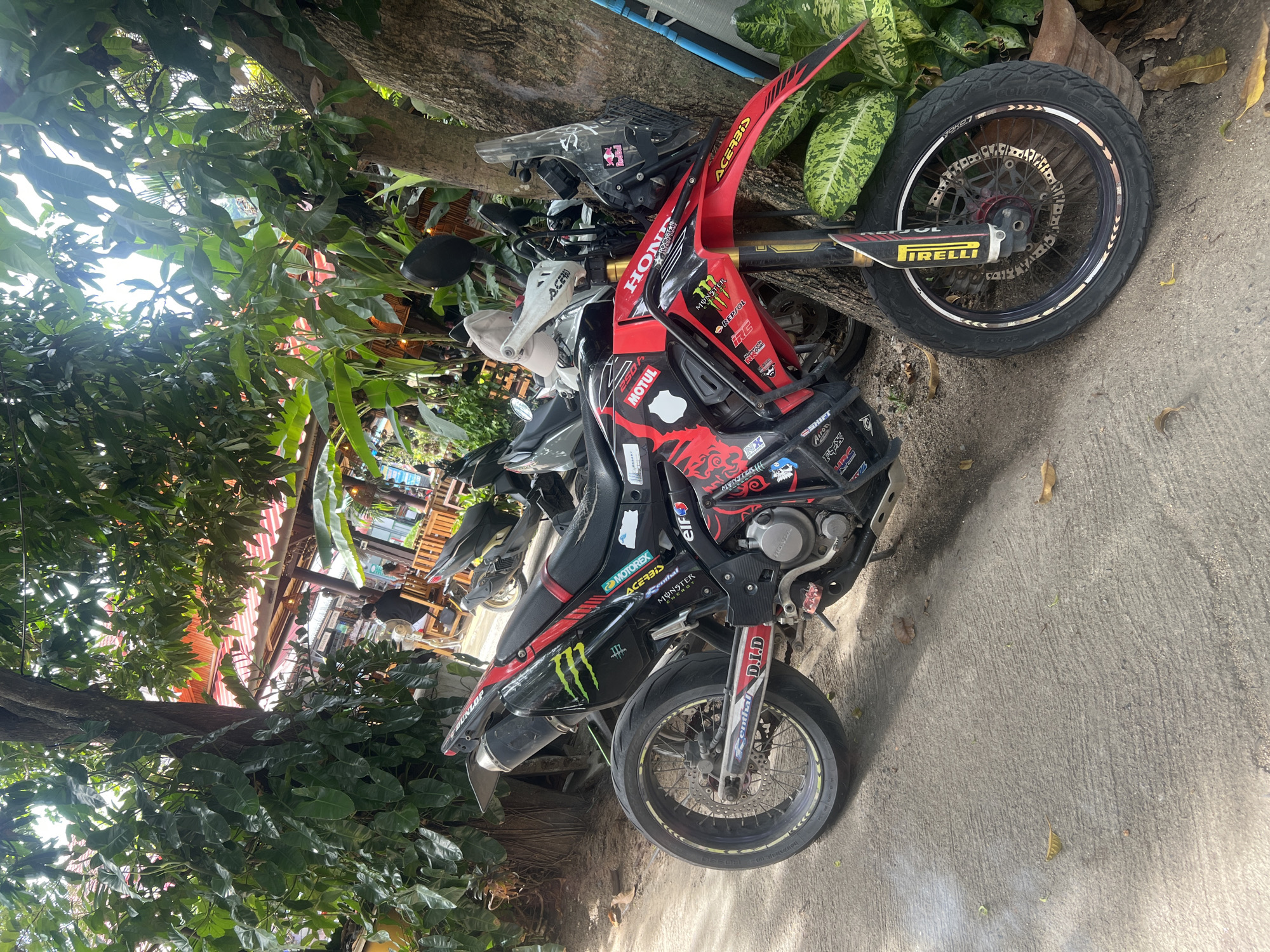 The image depicts a Honda 250A motorcycle, heavily adorned with racing-related stickers such as Acerbis, Motorex, Monster, Elf, and Pirelli, parked near a tree. A helmet with a Red Bull logo sits on the bike. The bike is parked on a concrete surface, adjacent to a leafy area. In the background, a building with an orange awning is visible, and people are seen sitting at tables. The location is identified as Baan Lamai, Thailand.