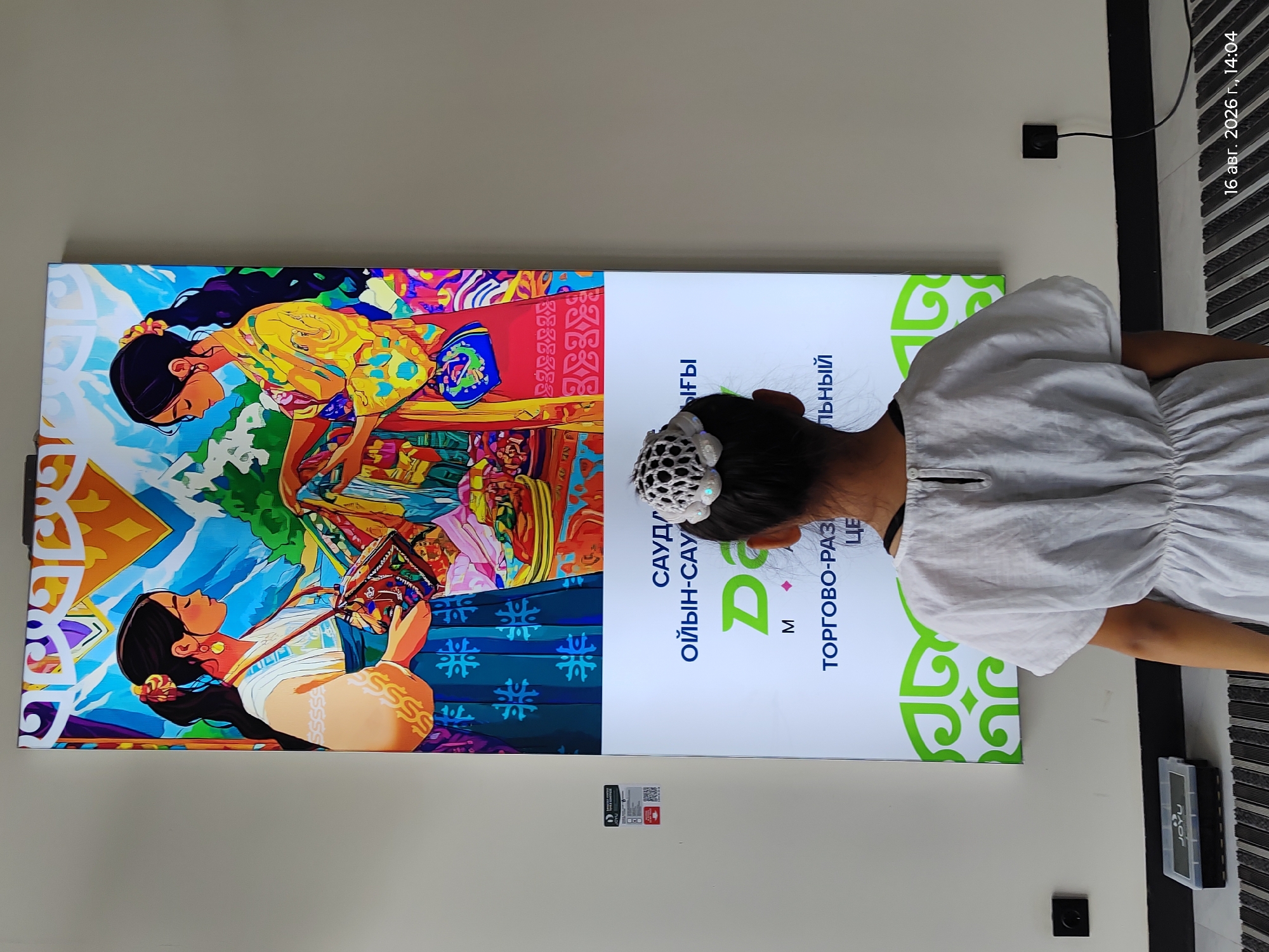 The image shows a young girl from behind, wearing a white dress and a decorative white hairpiece, looking at a large digital display. The display features an illustration of two young women in traditional attire and text in Cyrillic and Latin scripts. The background is a plain wall with a black baseboard. There are two electrical outlets on the wall, one with a black cord plugged in. On the floor in front of the display, there is a black object with a clear lid, labeled "JOYU". The image is dated "16 авг. 2026 г., 14:04" which translates to "August 16, 2026, 14:04". The text on the display includes the words "САУДА", "ОЙЫН-САУЫҚ", "M", and "ТОРГОВО-РАЗВЛЕКАТЕЛЬНЫЙ ЦЕНТР". The style of the illustration and the presence of Kazakh language elements suggest a location in Kazakhstan, likely Astana, given the Cyrillic script used. This could be an advertisement or information display within a shopping or entertainment center.