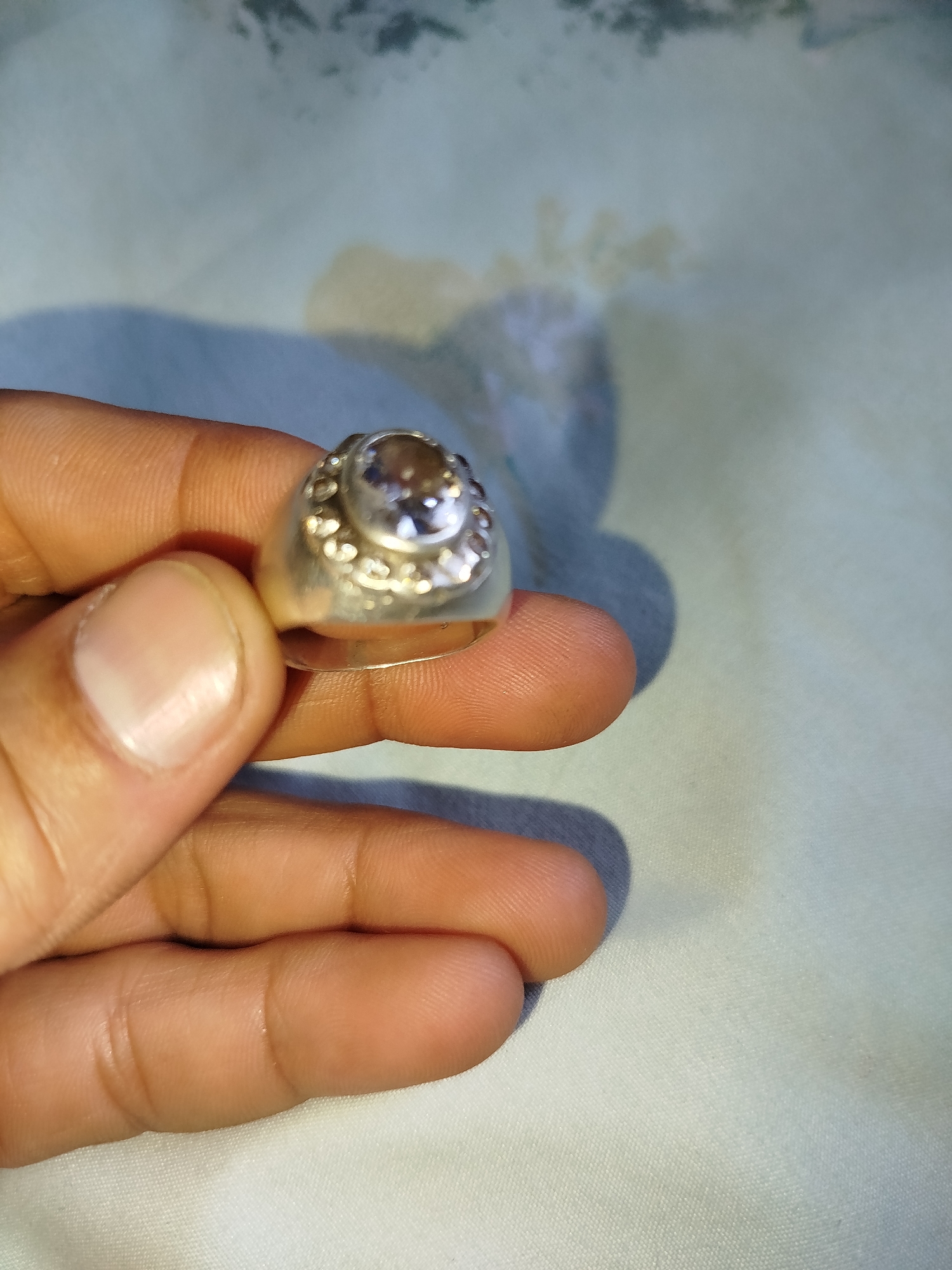In Kokand City, Uzbekistan, a person's hand is holding a silver ring with a clear gemstone. The ring is adorned with small circular embellishments around the gemstone, which is oval-shaped and appears to be faceted. The hand is positioned against a light-colored, textured fabric that could be a tablecloth or bedsheet. The lighting suggests an indoor environment, with shadows cast on the fabric by the hand and ring.