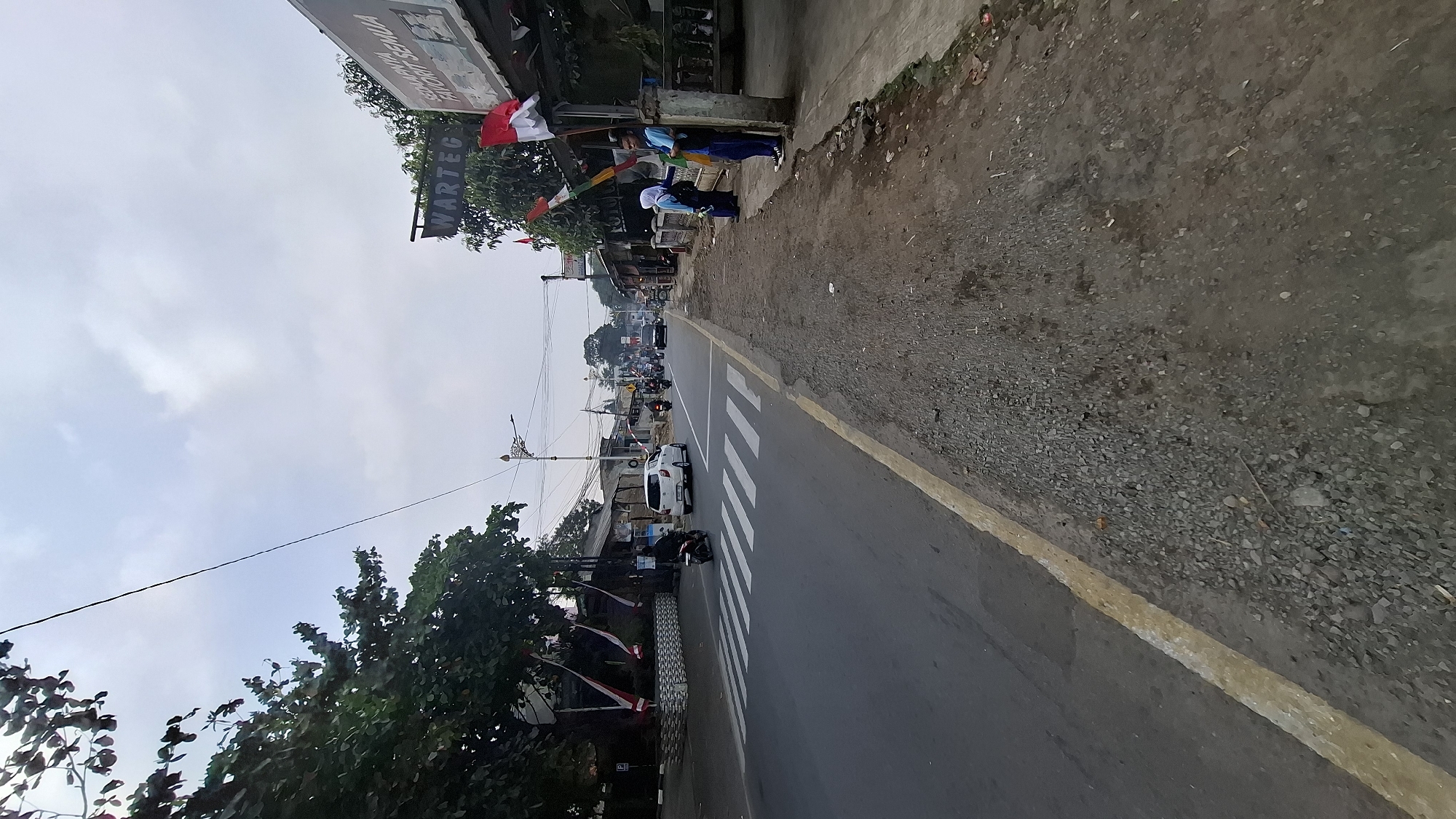 This image depicts a street scene in Cilaku, Indonesia. A paved road runs horizontally across the frame, marked with a yellow line on the right edge and a pedestrian crossing in the foreground. To the left of the road, there is a sidewalk area with a tree and several buildings. To the right, the sidewalk is unpaved and uneven, with some debris on the ground. In the background, there are more buildings and trees, with a cloudy sky overhead.
Several vehicles are present on the road, including a white car, a black motorcycle, and other vehicles further down the road. On the right sidewalk, two young individuals, likely students, are standing. One is wearing a light blue shirt and dark pants, and the other is wearing a light blue headscarf and dark clothing. Indonesian flags are visible, some hanging from poles and buildings, suggesting a local celebration or patriotic display. A sign with "WARTEG" is visible on one of the buildings. The environment appears to be a typical urban or semi-urban streetscape in Indonesia.
