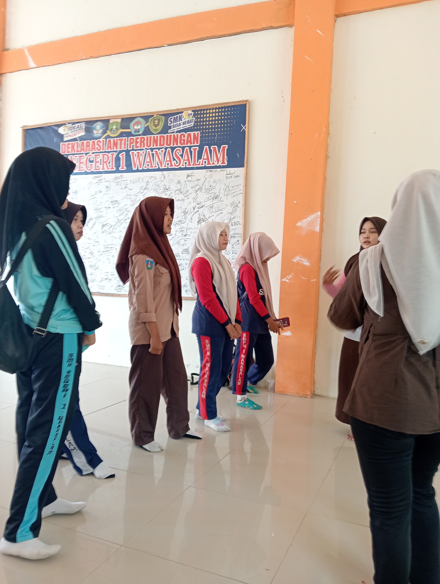 In a classroom or hallway in Wanasalam, Indonesia, several female students are gathered in front of a bulletin board. The bulletin board displays a blue banner with the text "DEKLARASI ANTI PERUNDUNGAN" (Anti-Bullying Declaration) and "NEGERI 1 WANASALAM" (Wanasalam State 1). The board is covered with handwritten signatures and messages. The students are wearing hijabs and uniforms, suggesting a school setting. One student in the foreground on the left is wearing a black and teal tracksuit and a black hijab, with a black bag slung over her shoulder. To her right, another student in a brown hijab and khaki uniform stands facing the bulletin board. Further to the right, two students in athletic wear, one with a pink and blue top and the other with a red and blue top, also appear to be looking at the board or interacting with each other. A student on the far right, wearing a brown hijab and a brown jacket over dark pants, is gesturing with her hands while seemingly talking to another student whose face is obscured. The environment appears to be indoors with tiled flooring and a light-colored wall with an orange structural beam or column. The overall scene suggests a school activity or event related to anti-bullying.
