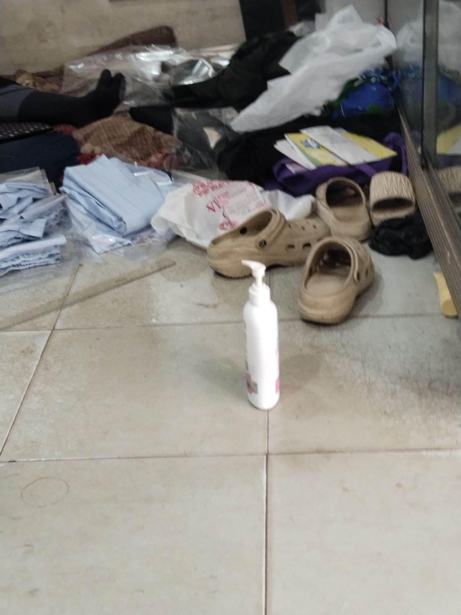 The image depicts a cluttered indoor scene, likely a storage area or a room with disarrayed belongings. The floor is tiled with light-colored square tiles. In the foreground, a white plastic bottle with a pump dispenser is standing upright on the floor. Next to it are several pairs of tan colored clogs. In the background, there are piles of folded light blue fabric, some plastic-wrapped items, and other assorted objects suggesting untidiness. A person's legs, wearing black split-toe socks, are visible on the left side of the image, extending from beneath a dark fabric. The lighting is dim, and the overall atmosphere is one of disorganization. The specific location within Unknown, Indonesia cannot be determined from the visual information provided.