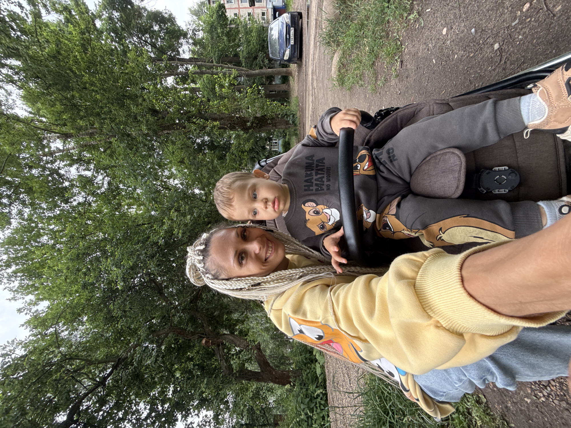 In Rusanovo, Russia, a young woman with blonde braided hair smiles as she takes a selfie outdoors. She is wearing a yellow hoodie with a cartoon character on the sleeve. Beside her, a young child sits in a stroller, dressed in a grey sweatshirt and pants featuring characters from The Lion King, including the phrase "Hakuna Matata." The stroller has a black handlebar. The background shows a dirt path, green trees, and a building with windows. A dark-colored car is parked on the right side of the frame.