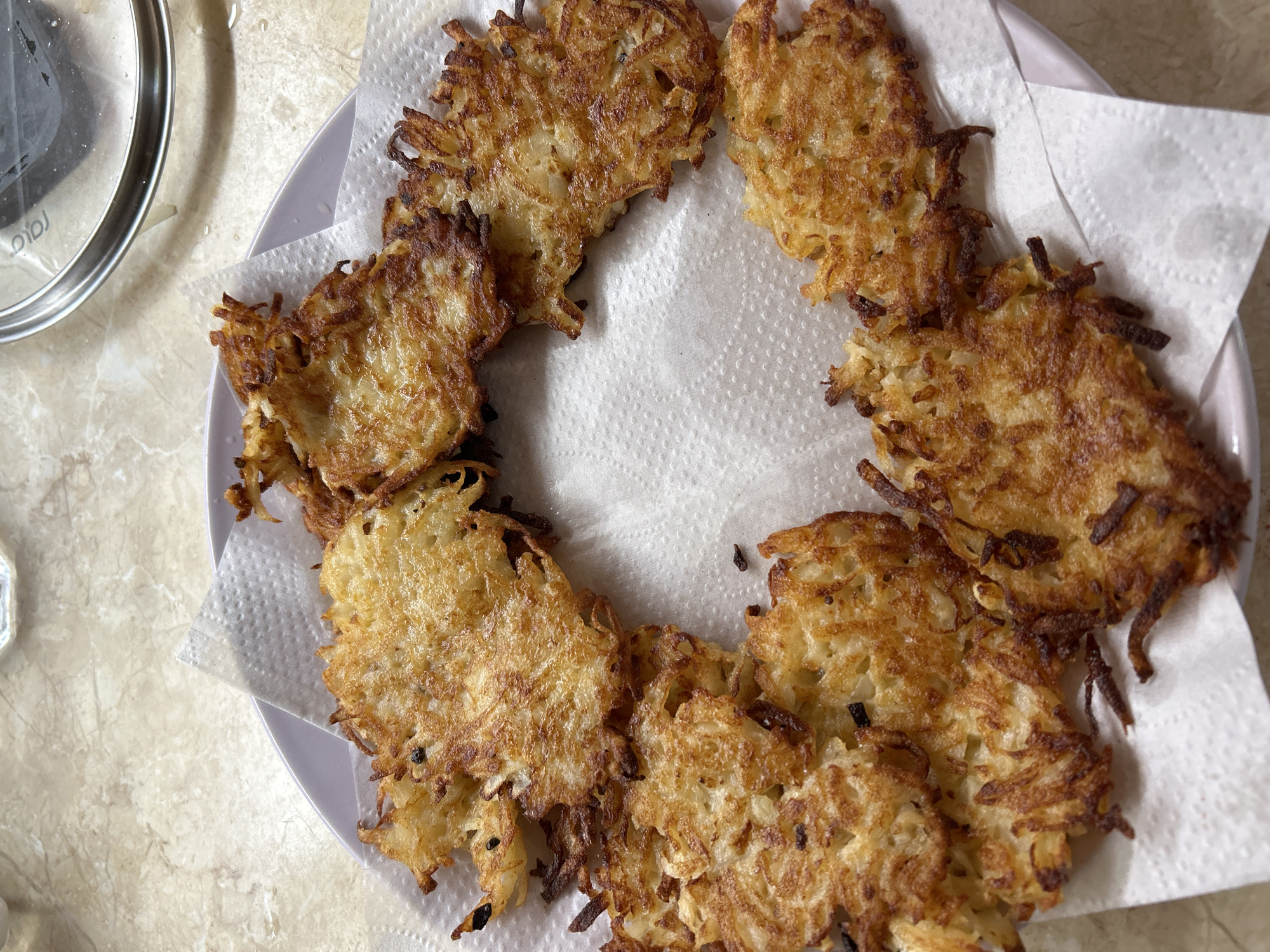 A top-down, close-up, high-angle shot shows a plate of golden-brown potato pancakes arranged in a circular pattern on top of white paper napkins. The potato pancakes are thin and crispy, with visible shredded potato strands. Some of the edges of the pancakes are darker brown and appear fried. The background is a light-colored, speckled countertop. A portion of a metal pot lid is visible in the upper right corner of the image. The scene appears to be in a kitchen setting, though the specific city and state are not identifiable from the visual information.