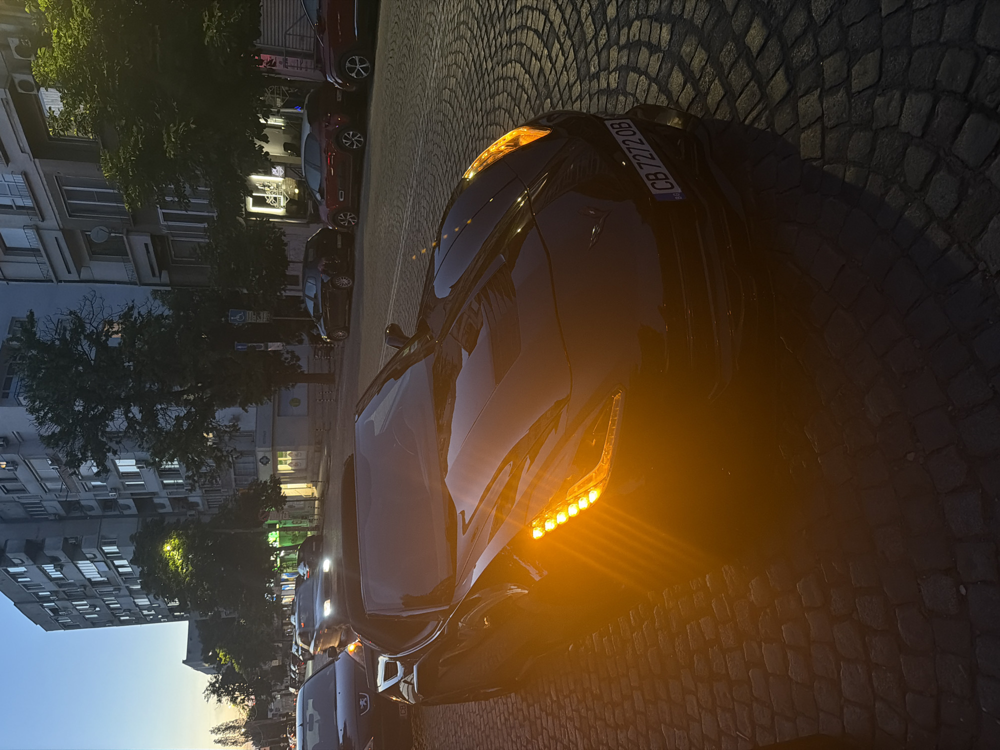 The image depicts a street scene at dusk in Sofia, Bulgaria. A black convertible sports car with its hazard lights illuminated occupies the foreground, parked on a cobblestone street. Behind the sports car, other vehicles are visible, suggesting moderate traffic. The background features a multi-story apartment building with numerous windows, some illuminated. Trees line the street, and streetlights cast a warm glow. The sky is a deep blue, indicative of twilight. The license plate of the sports car is partially visible, reading "CB 7272 OB". The overall atmosphere is urban and calm, with the setting sun or artificial lights creating a dramatic interplay of light and shadow.