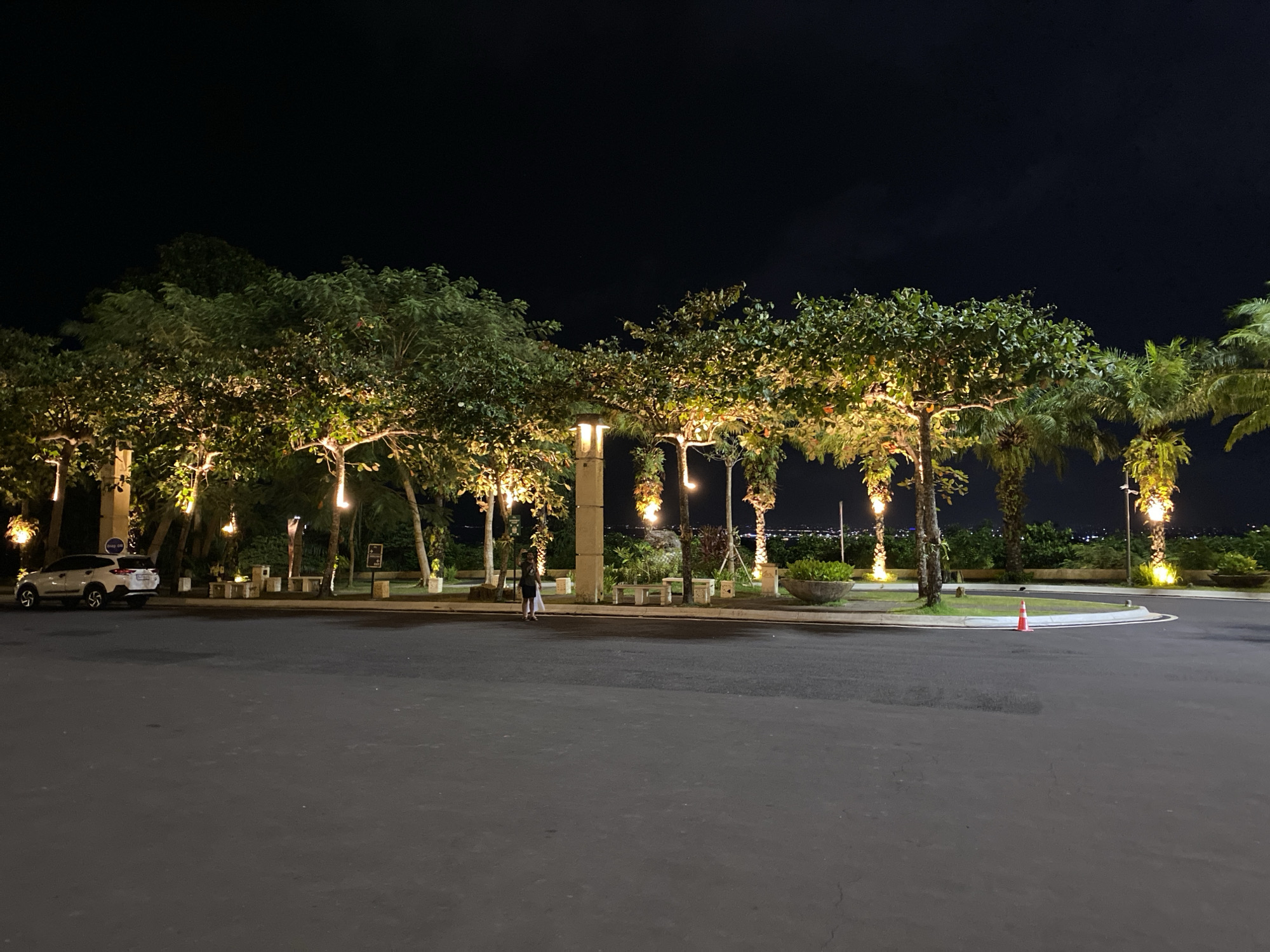An outdoor wide shot, taken at night, captures a paved area with a white SUV parked to the left. The SUV is facing the right side of the frame. Further into the scene, there are several trees, some with illuminated branches, lining a pathway. A lone person, appearing to be walking, is visible in the middle ground. In the background, there is a subtle, distant city glow. The overall atmosphere is serene and dimly lit.
