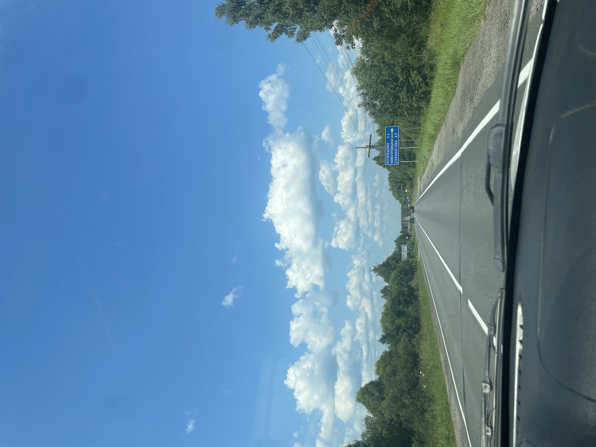 The image shows a view from inside a car, looking out at a highway on a sunny day. The sky is bright blue with fluffy white clouds. On the right side of the road, there is a blue directional sign with white text in Russian. The sign indicates "КУЗНЕЦОВО 2.2", "НОВОТРОИЦКОЕ 2.9", and "СУХОНОГОВО 3.3". This suggests the location is near Yasiplevo, Russia, as it is a known road leading to these locations. There are trees and greenery lining both sides of the road, and a few other cars are visible in the distance. The dashboard of the car is visible in the foreground.