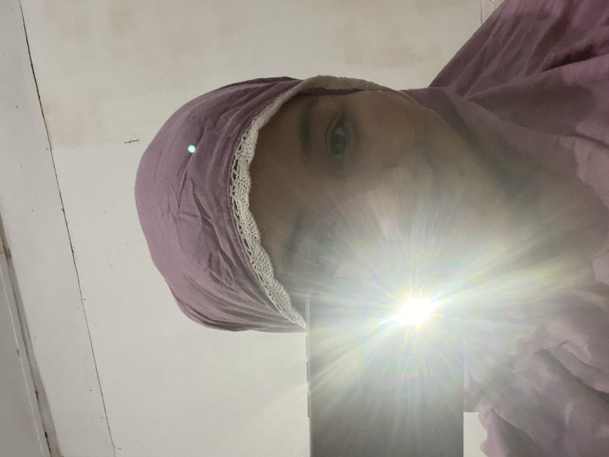 The image shows a close-up, low-angle shot of a person's face, mostly obscured by a purple headscarf with a lace trim. A bright light source, possibly a camera flash or phone light, is pointed directly at the camera, creating a strong lens flare that obscures much of the person's features. One eye is partially visible. The background appears to be a plain wall, possibly with some subtle texture or dirt marks. The lighting suggests an indoor setting. The composition and the bright light indicate that this is likely a selfie, taken in Banjarbaru, Indonesia. No text is visible in the image. The overall impression is informal and personal, with the bright light adding a dynamic, almost dramatic effect to the otherwise simple composition.