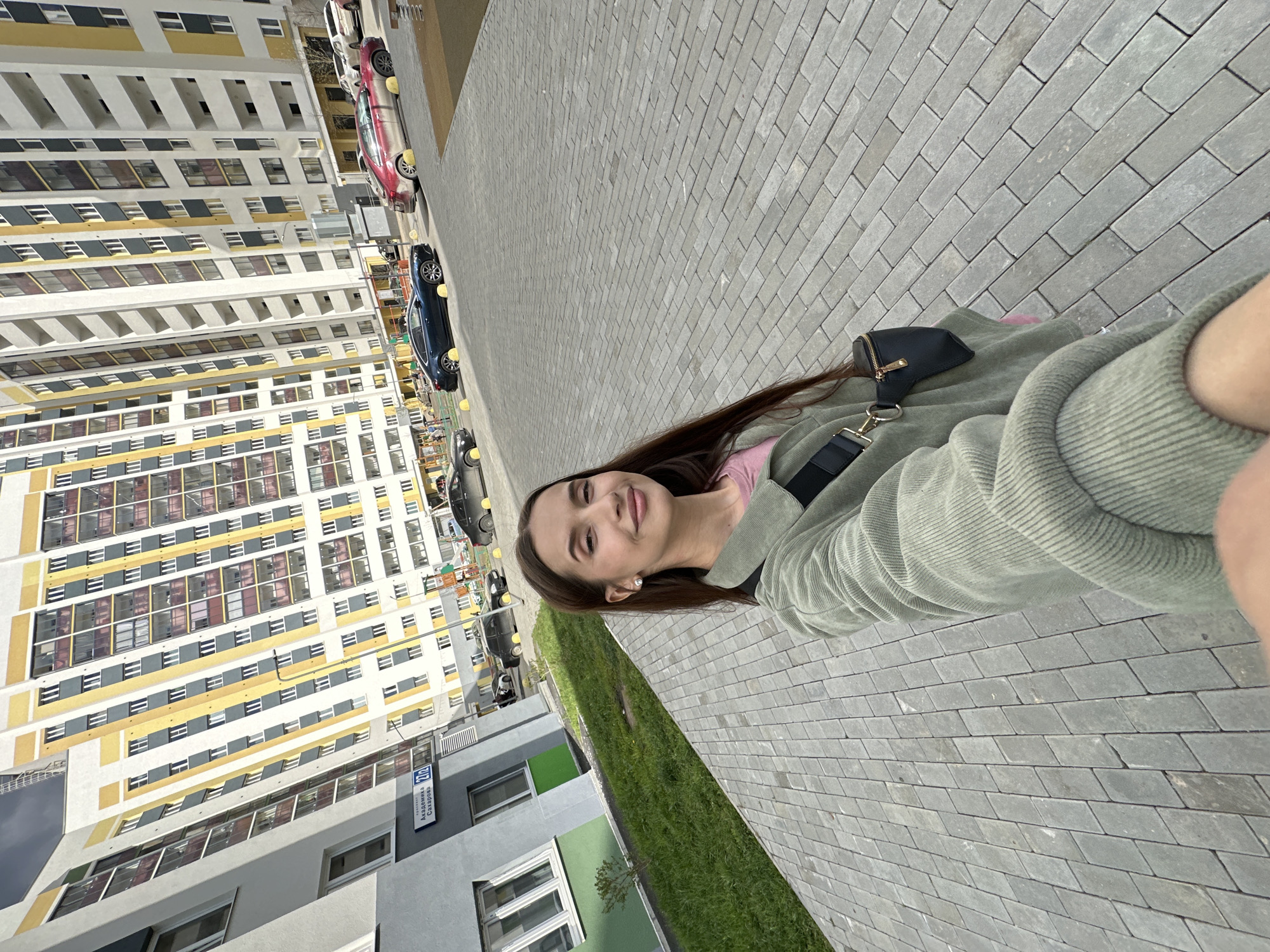 A woman is standing on a gray cobblestone walkway in Yekaterinburg, Russia, presumably taking a selfie. The background shows a tall residential building with a repeating pattern of windows and yellow and white panels. Various cars are parked along the street in front of the building. A strip of green grass separates the sidewalk from a lower building with gray walls and green accents. The woman is wearing a light green jacket and a small black purse with a shoulder strap.