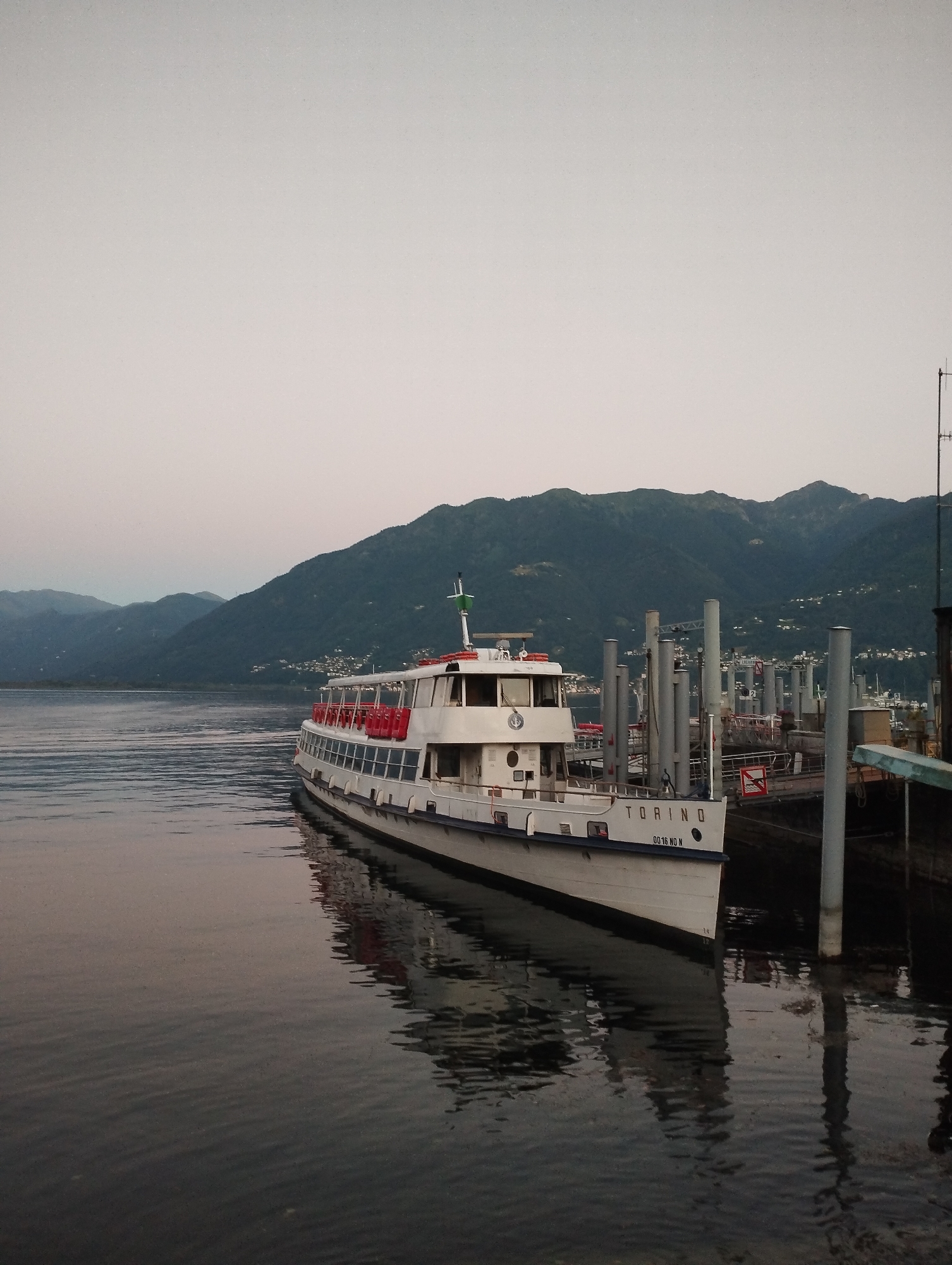 A wide shot captures a ferry named "TORINO" docked in the calm waters of a lake, with a backdrop of dark, tree-covered mountains. The sky is a soft, overcast gray, and the water reflects the ferry and the muted colors of the landscape. The ferry, predominantly white with red accents on its upper deck railings, is positioned parallel to a pier structure. The pier is characterized by several tall, cylindrical grey posts and some docked boats visible in the background to the right. The "TORINO" text is clearly visible on the side of the ferry. The scene appears to be set in Muralto, Switzerland, a location known for its lakefront and surrounding mountains. The image is captured at what appears to be either dawn or dusk, given the soft lighting and the lack of harsh shadows. There are no people visible in the frame.