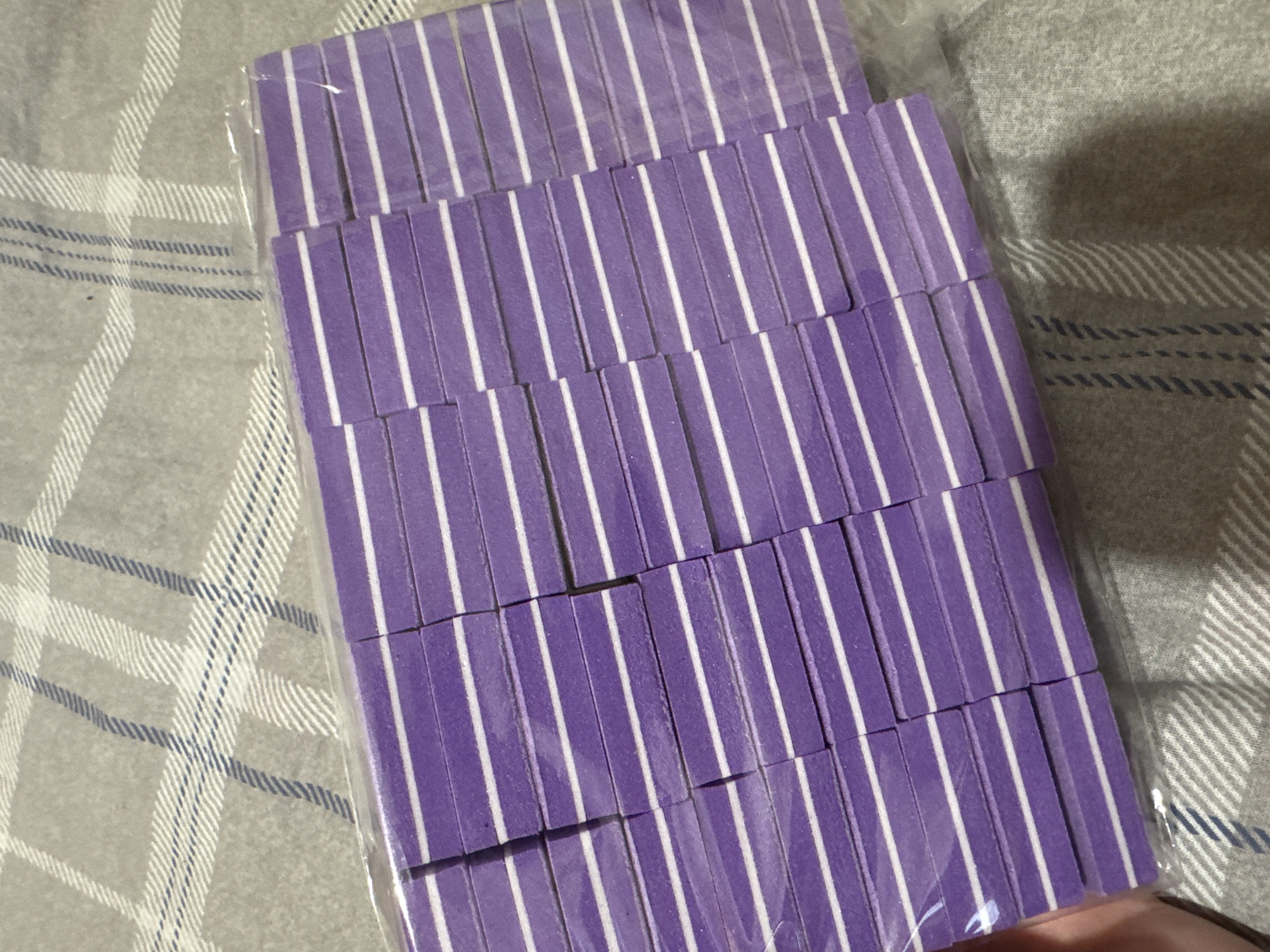 This media file shows a top-down view of a package of purple and white striped nail files. The nail files are arranged in neat rows within a clear plastic wrapper. The background consists of a gray fabric with a plaid pattern, featuring lines in white and dark blue. No people or activities are depicted. The lighting suggests an indoor setting, possibly a home environment. There is no visible text in the image. The context provided indicates the location as Yarominski Village Council, Belarus.