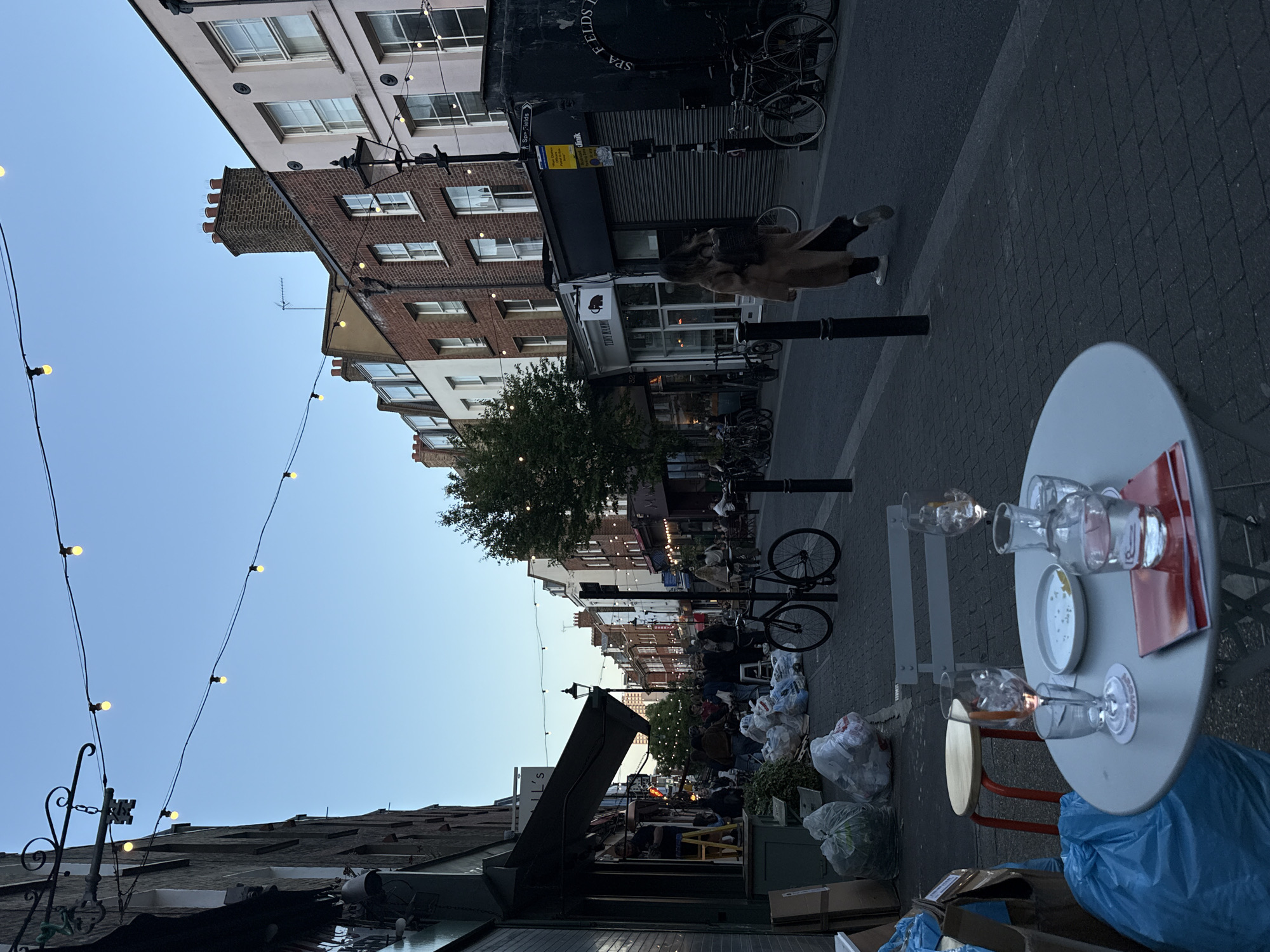 The image depicts a quiet urban street scene, likely in the early evening or at dusk, given the soft, fading light in the clear blue sky and the illuminated string lights overhead. The street is lined with multi-story buildings, featuring traditional brickwork and painted facades with numerous windows, characteristic of older European architecture.
In the foreground, a white, round outdoor table is visible on the right, set with several empty glasses and a small plate containing crumbs, suggesting recent use. A red menu or paper lies on the table, and a blue trash bag rests beneath it. To the left of the table, various white and blue trash bags are piled on the sidewalk, indicating a cleanup or end-of-day activity, possibly from nearby shops or market stalls further down the street.
A lone person, dressed in a light brown coat, walks in the mid-ground, moving away from the viewer. Several bicycles are parked along the street, leaning against poles or racks. As the street recedes into the background, more buildings, trees, and indistinct figures can be seen.
Notable details include string lights with yellow bulbs strung across the street, adding a festive touch. On one building, the partial text "SPA FIELDS" and "KLINIK" are visible on a dark facade, with an arrow on a street sign pointing towards "Spa Fields." Another shop sign displays a black cat or animal logo with the text "TINY HANDS." Chimney pots adorn the rooftops of some buildings, and a television antenna is also visible against the sky. The overall atmosphere is tranquil and somewhat melancholic, as the day transitions to night.
