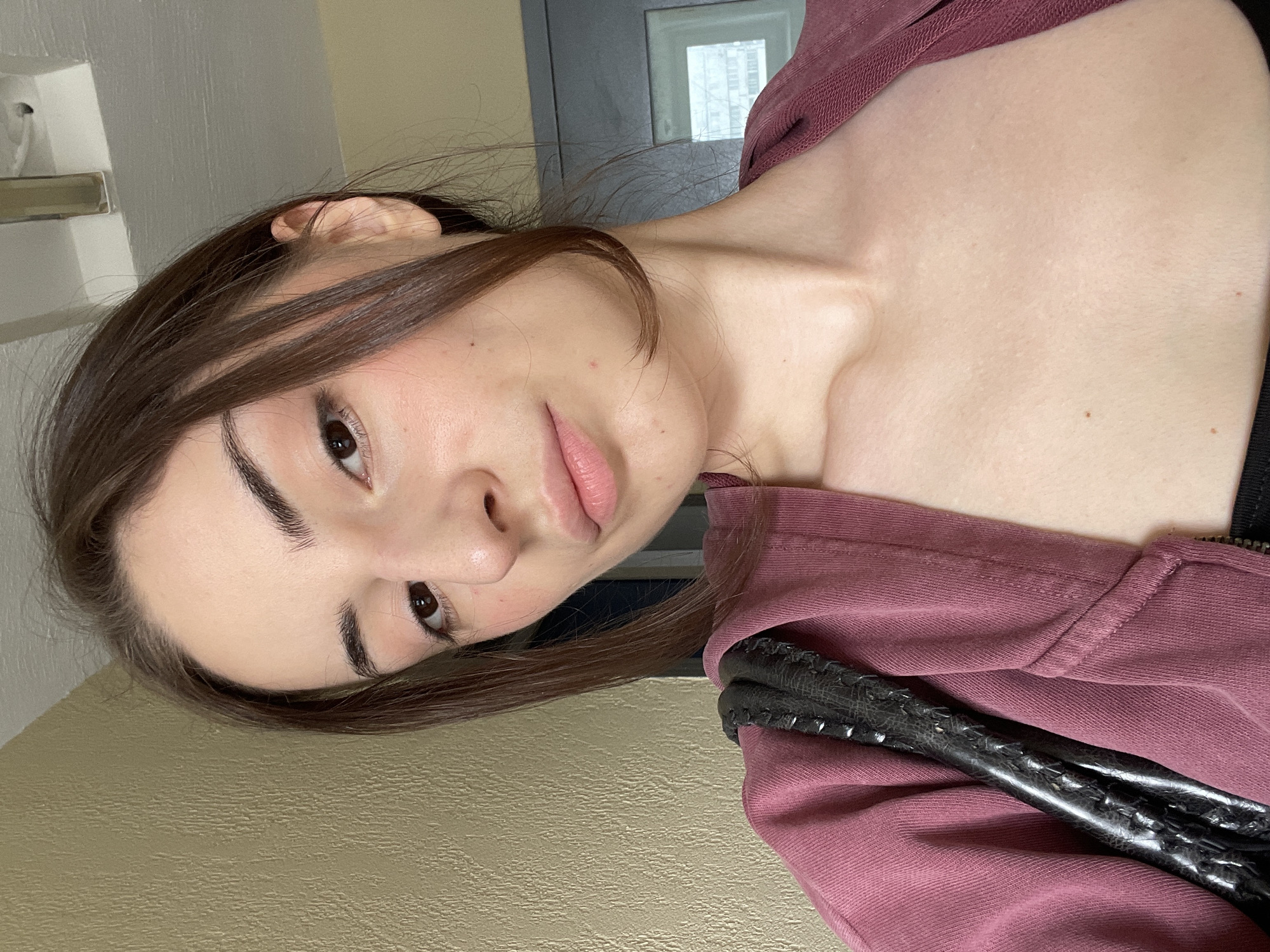 A young woman with medium brown hair and brown eyes is pictured in a close-up, head-and-shoulders shot. She is wearing a mauve hooded jacket and a black top. A textured black handbag strap is visible on her left shoulder. The background consists of a textured yellowish wall on the left and a doorway with a window on the right. The scene is indoors, likely an apartment or hallway, with no discernible Moscow landmarks.