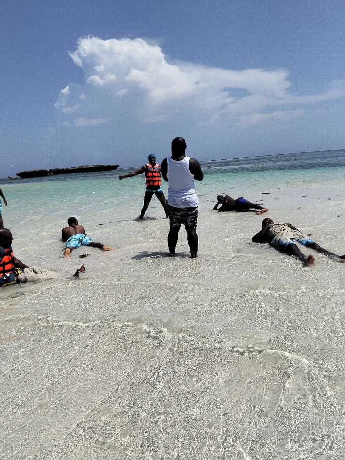 This image depicts an outdoor scene in Wasini, Kenya, on a clear day with a blue sky and a large white cloud in the upper left. The foreground and midground consist of shallow, transparent turquoise ocean water revealing a light-colored sandy seabed with ripples. Further back, the ocean deepens to a darker blue, with a low-lying, dark, elongated object (possibly a rock formation or landmass) visible in the distant water to the left.
Six individuals, all appearing to be adult males with dark complexions, are present in the shallow water.
1. A man stands in the central midground with his back to the viewer, wearing a white tank top and dark, patterned shorts.
2. To his left, another man lies prone on his stomach in the water, wearing light blue and white patterned swim trunks.
3. Further left, a man is seated on a small sandy mound partially submerged in the water, wearing an orange life vest over a dark shirt and dark shorts. A small reddish-brown object is near him in the water.
4. In the midground-background, another man stands facing away from the camera with his arms slightly outstretched, wearing an orange life vest, dark swim trunks, and a blue head covering or cap.
5. To the right of the central standing man, a fifth man lies prone on his stomach in the shallow water, wearing dark blue swim trunks.
6. The sixth man lies supine on his back in the shallow water towards the right side of the frame, wearing light-colored patterned swim trunks.
The individuals are positioned in various states of rest or relaxation within the calm, clear water.