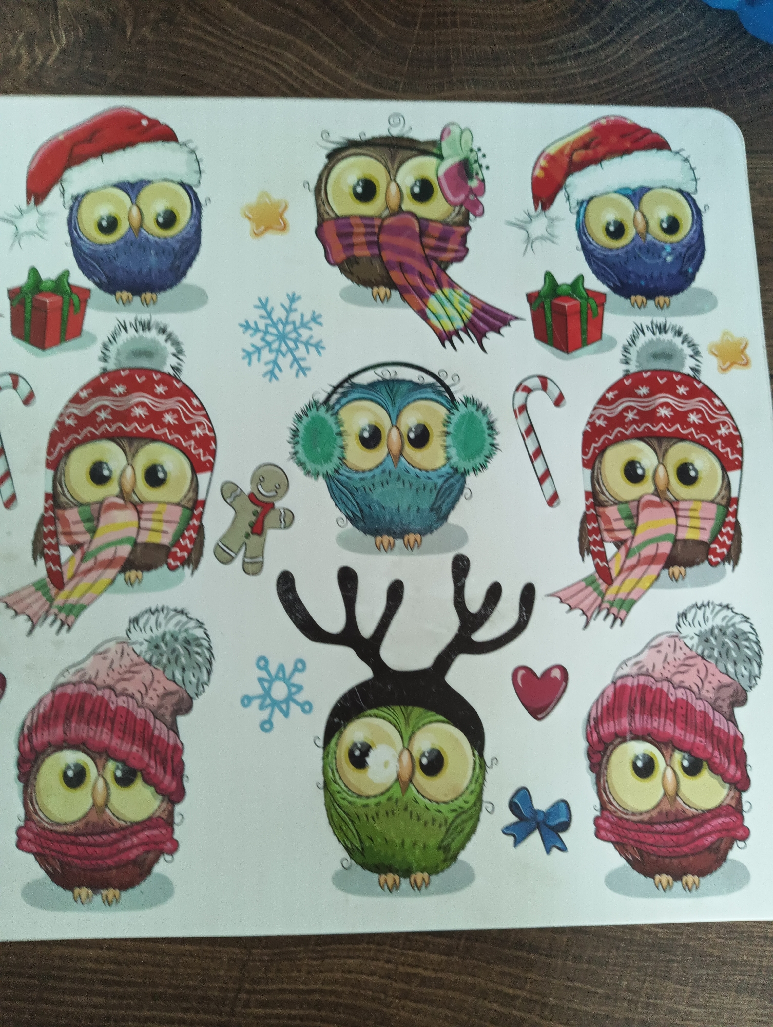 The image displays a collection of cartoon owls, rendered in a festive, winter-themed style, arranged on a white surface with a visible wood grain texture in the background. There are nine owls in total, each with large, expressive eyes and adorned with winter accessories such as Santa hats, scarves, and earmuffs. Some owls are accompanied by small, decorative elements like gift boxes, snowflakes, stars, a candy cane, a gingerbread man, antlers, a heart, and a blue bow. The owls vary in color, with some being brown, blue, and green. The overall presentation suggests a decorative item or a sheet of stickers. The location is not discernible from the image.