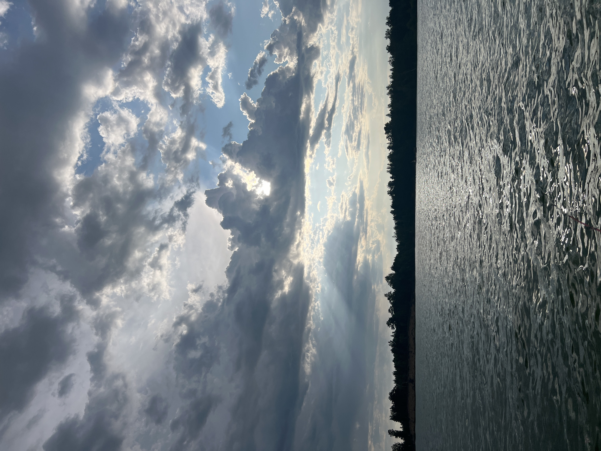 The image depicts a serene lake scene with a dramatic sky. The lake's surface is textured with ripples and reflects the light in shimmering patterns. A dense line of dark green trees forms a silhouette along the distant shore. Above the trees, a vast sky is filled with a dynamic arrangement of clouds. Some clouds are dark and heavy, suggesting an impending storm, while others are lighter, allowing shafts of sunlight to break through, illuminating the scene with a dramatic effect. The light filtering through the clouds creates a sense of depth and highlights the edges of the cloud formations. The overall impression is one of natural beauty and the powerful, ever-changing moods of the sky. The scene is set in Borovoe, Russia, a location known for its picturesque lakes and surrounding forests. There are no people or specific actions depicted in the image.