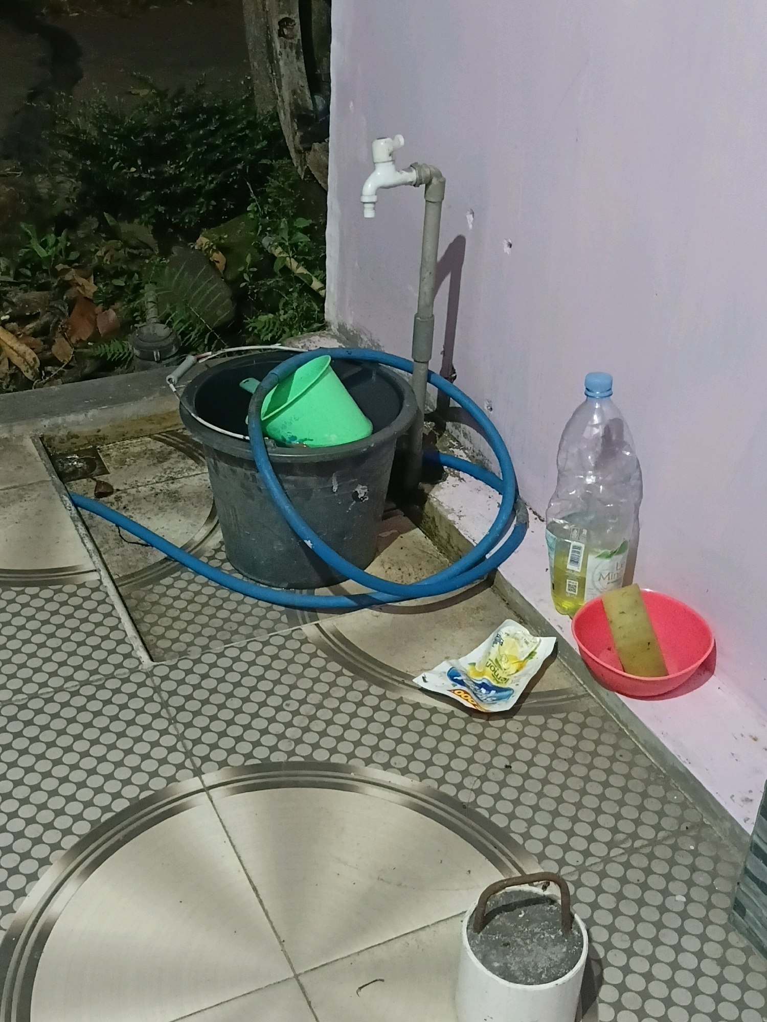 This is a low-angle, medium shot of an outdoor utility area at night, possibly in Cikal, Indonesia. The scene features a grey bucket with a green funnel inside, a blue hose coiled around the bucket, and a white faucet attached to a grey pipe against a light purple wall. To the right of the faucet, there is a large, empty plastic water bottle with a blue cap, and next to it, a pink bowl containing a yellow sponge. On the tiled ground in front of the bottle and bowl, there is an opened packet of what appears to be cleaning wipes. In the foreground, a white cylindrical object with a rusty metal handle is visible on the patterned tiled floor. In the background, lush green foliage and some dried leaves are present, suggesting an outdoor, possibly garden, setting. The lighting is dim, indicative of nighttime.