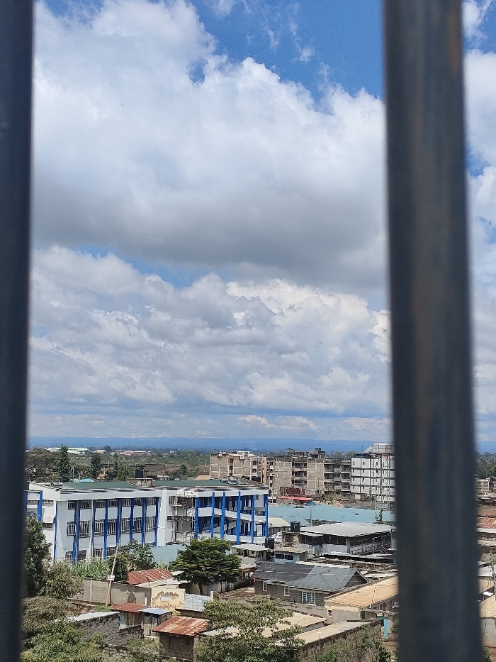 The image presents an elevated, wide-angle view of an urban landscape within Juja ward, Kenya, framed by two prominent, out-of-focus vertical metallic bars on the left and right edges. The upper two-thirds of the scene depict a partly cloudy blue sky, featuring large, white cumulus clouds. Below the sky, the horizon reveals a distant, hazy landmass, possibly a mountain range or hills.
The midground is dominated by a dense arrangement of buildings. A distinctive multi-story building with a white exterior and vertical blue architectural accents is visible on the mid-left. To its right, and extending into the background, are numerous other multi-story structures, several of which appear to be under construction, characterized by exposed concrete frames and rebar. The lower midground consists of closely packed smaller residential and commercial buildings, many featuring corrugated iron roofs, some with visible signs of rust, and others with flat roofs often supporting water tanks. Interspersed throughout the built environment are various green trees and patches of foliage. No individuals or specific activities are discernible within the frame.