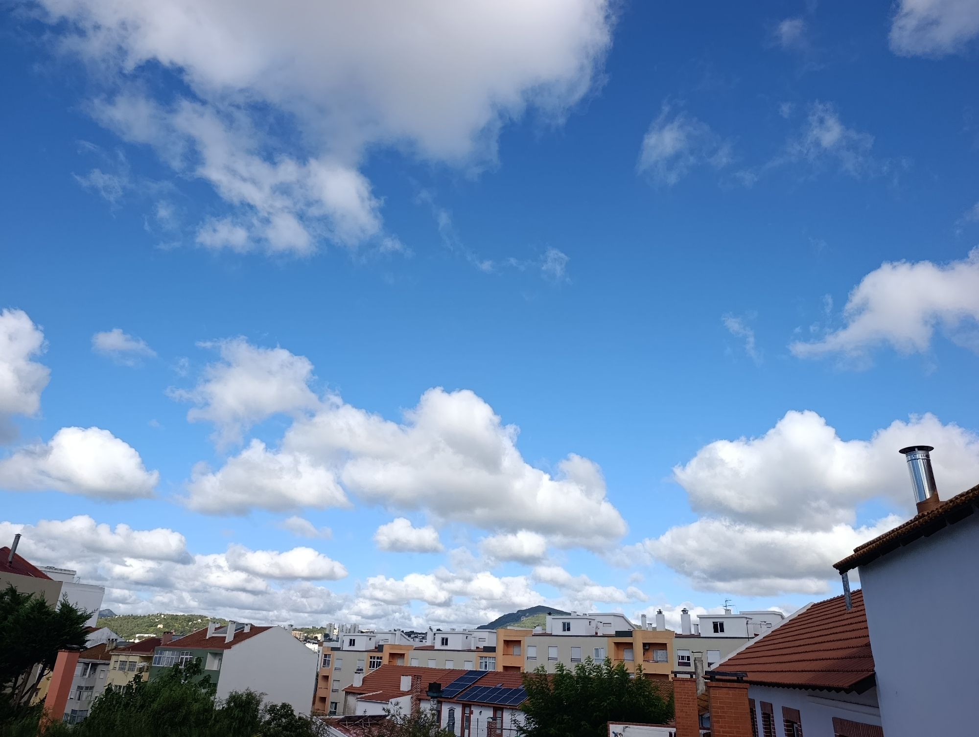 This is an eye-level, medium shot photograph taken on a sunny day, featuring a residential cityscape under a bright blue sky dotted with fluffy white clouds. The foreground is dominated by the rooftops of several buildings. On the right, a white building with a brown tiled roof is visible, featuring a prominent stainless steel chimney. To its left, a closer view of buildings shows red tiled roofs, some with visible solar panels. Further into the background, a range of apartment buildings with neutral colored facades and white window frames are apparent. Some buildings have balconies. In the distance, a rolling, green hillside is visible, with a mountain peak in the far background. The image depicts a typical urban residential area. The photo was taken in the city of Setúbal, Portugal.
