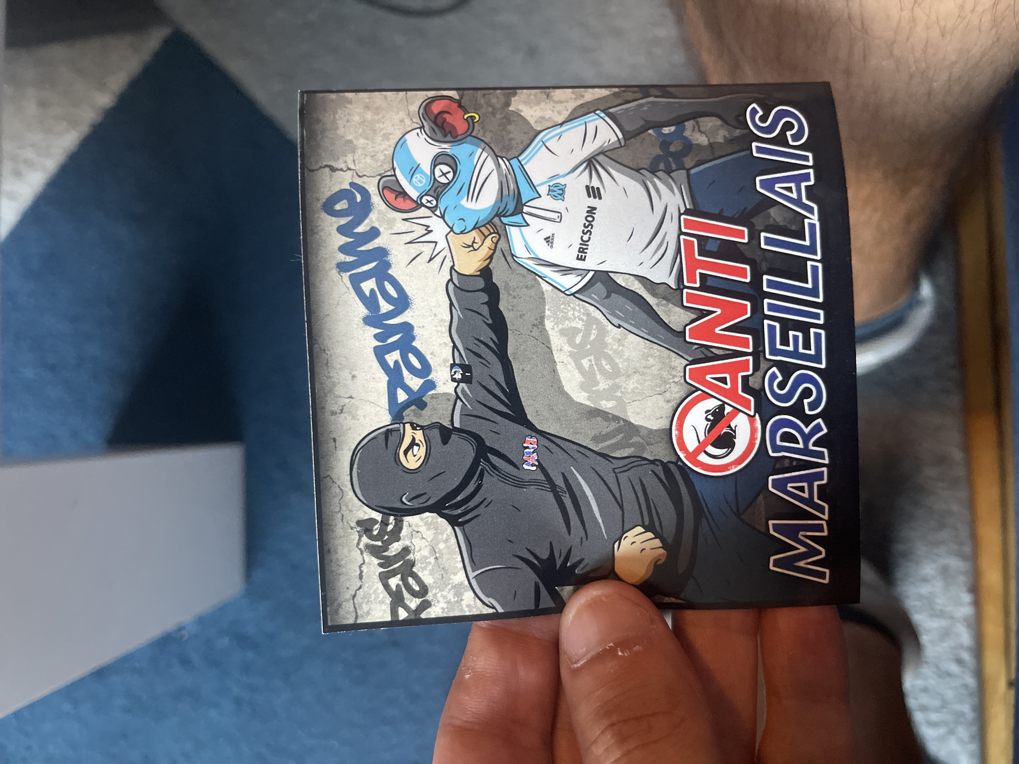 A sticker features a cartoon drawing of two figures. On the left, a person wearing a black balaclava and grey tracksuit punches a cartoon mouse on the right. The mouse wears a blue and white racing helmet with red ears, and a white polo shirt with blue stripes and logos that appear to be for Adidas and Ericsson. The background is a grey, cracked wall with graffiti. Above the figures, blue graffiti reads "Parame." Below the figures, red text reads "ANTI" inside a white circle with a red border, and blue text below that reads "MARSEILLAIS". The sticker is held in a person's hand, with a blue carpeted surface and a white object visible in the background. The person's hand is holding the sticker at an angle, with their thumb and index finger on the left side. The sticker appears to be a political or sports-related message, likely referencing the rivalry between different groups or sports teams. The location in Paris, France is not discernible from the image.