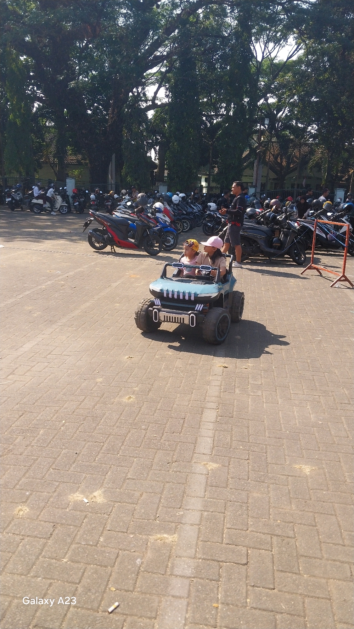 In Malang, Indonesia, on a sunlit day, a light blue toy jeep with two young children inside is parked on a paved area. The children, one wearing a yellow cap and the other a pink cap, are seated in the jeep. In the background, a man in a dark shirt and shorts stands with a group of motorcycles and scooters. Tall trees with dense foliage line the area, casting shadows on the ground. The paved surface is made of interlocking bricks, and scattered on the ground are some dry grass and what appear to be cigarette butts. A red metal barrier with orange poles stands to the right of the motorcycles. The image is captured from a slightly elevated angle, looking down towards the toy jeep.