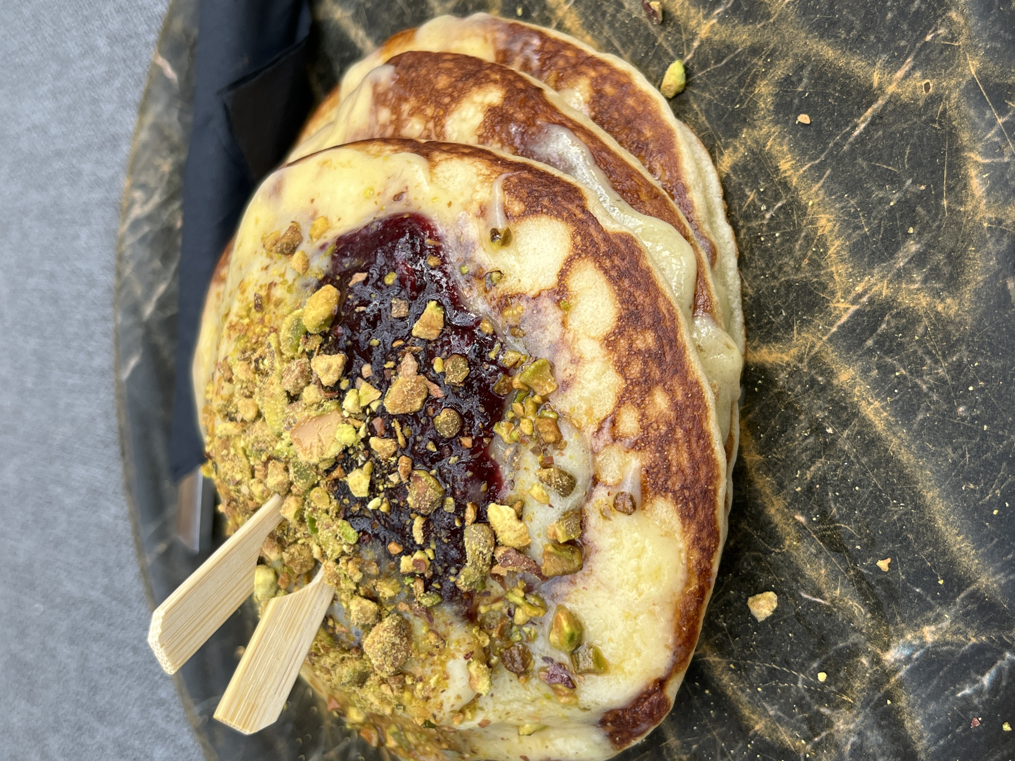 A stack of three golden-brown pancakes is presented on a dark, textured surface. The top pancake is topped with a dollop of dark red jam, and generously sprinkled with chopped pistachios and what appears to be crumbled biscuit or cookie pieces. Two small, pointed wooden skewers are inserted into the topping. The pancakes have a slightly glossy sheen, suggesting they may be lightly buttered or syrupy. The background is out of focus, showing a gray textured surface and a dark fabric. The scene is captured in a close-up, overhead shot. This image was taken in Thassos, Greece, at a restaurant called "The Garden."