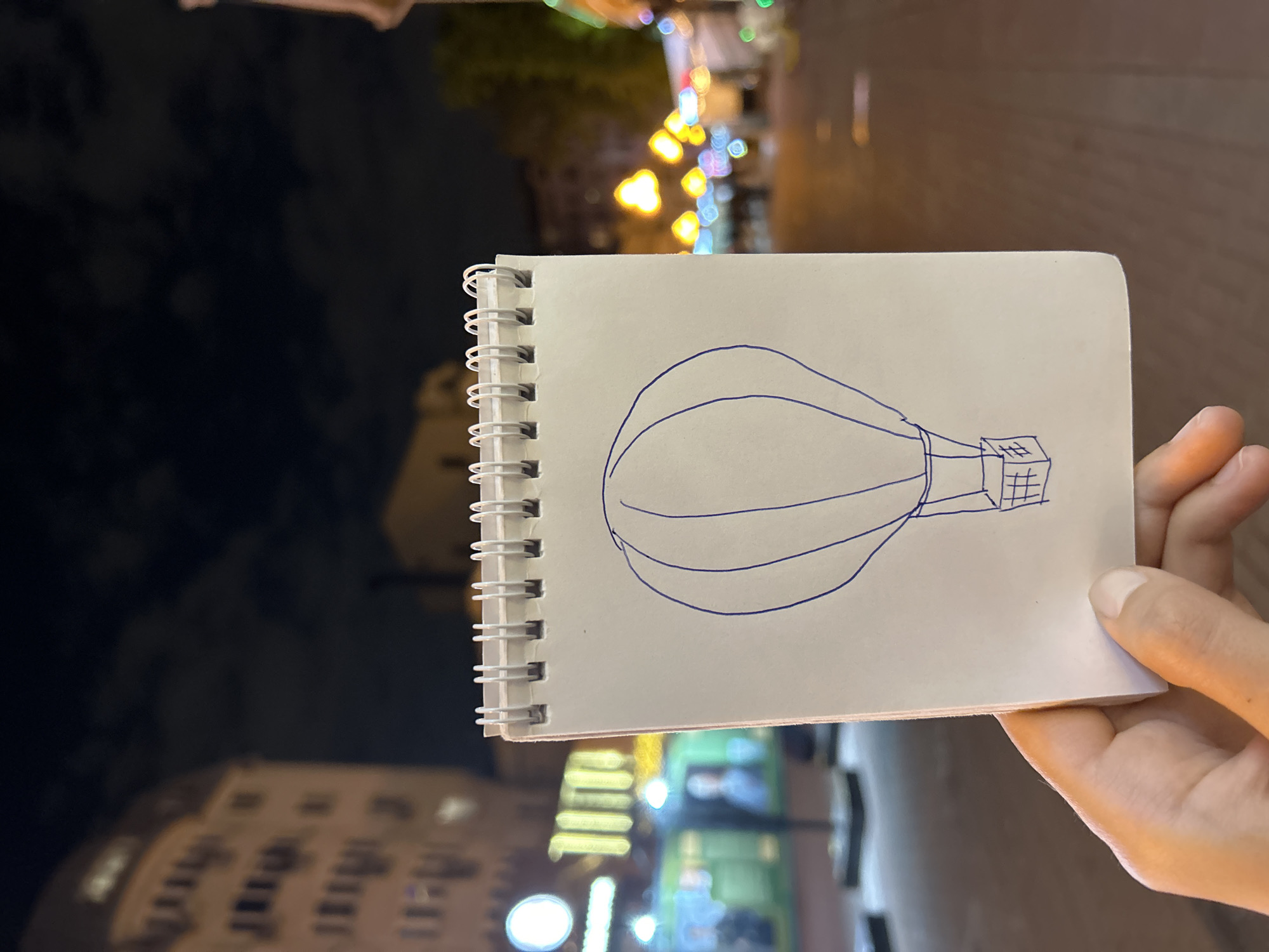 A person holds a notepad in front of an urban scene at night. The notepad contains a simple drawing of a hot air balloon in blue ink. Behind the notepad, blurred lights and buildings are visible, including string lights and building windows. The background is mostly dark. The hand holding the notepad is visible on the right side of the image.
