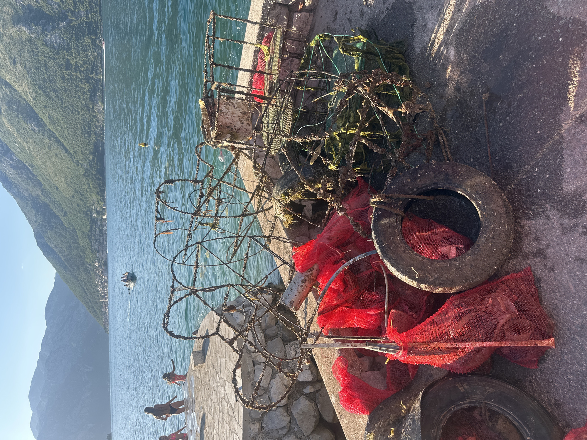 The image depicts a coastal scene in Prčanj, Montenegro, under clear skies with the sun low in the sky, casting shadows to the left.
In the immediate foreground, a concrete pier or embankment extends into a body of water. Piled on this concrete are several weathered and corroded items, including multiple fishing or crab traps constructed from rusted metal frames, some covered in marine growth. Interspersed with these traps are several discarded vehicle tires and multiple red mesh bags, some appearing to contain dark, indistinct objects. A corroded metal pipe or rod is also visible among the debris. The concrete surface is discolored and shows various stains.
In the midground, the calm, blue-green water of a bay extends from the pier. Four individuals, appearing to be young females, are present near the water's edge on the left. One person in a pink swimsuit is partially visible entering the water. Another, wearing a black bikini bottom, is stepping into the water. A third, in a patterned swimsuit, wades in the water, facing away from the camera. A fourth person is partially obscured behind the first. Further out in the bay, a small motorboat carrying multiple passengers is moving towards the left, leaving a wake behind it. A single yellow buoy floats in the water to the right of the boat.
The background is dominated by large, steep mountains covered in green vegetation, rising from the water's edge. On the opposite side of the bay, nestled at the base of the mountains, a cluster of buildings forms a distant settlement. The sky above is a clear, light blue.