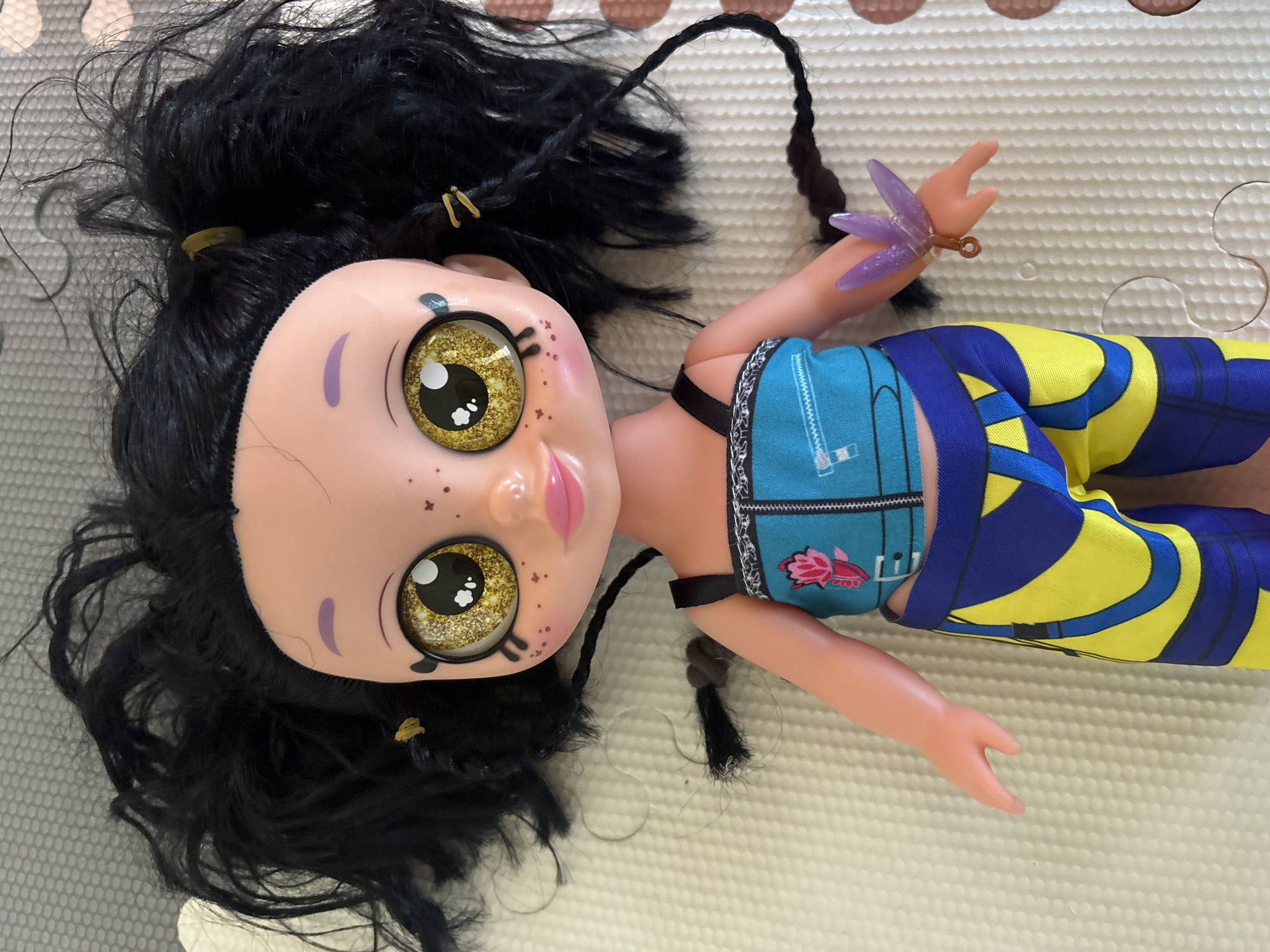 A light-skinned plastic doll is depicted lying supine on a light-colored, textured foam mat, situated in the city of Dubai, United Arab Emirates. The doll features prominent, glittery golden eyes with large black pupils and white catchlights, pink lips, small brown freckles across its cheeks, and thin purple eyebrows. Its black, synthetic hair is styled in a disheveled manner, with two small ponytails secured by yellow elastic bands and two partially unraveled braids; one long braid extends along its right side. The doll is clothed in a teal crop top with black shoulder straps, featuring a printed zipper detail and a pink lotus flower motif on the left chest. Its lower attire consists of blue and yellow patterned pants with abstract curvilinear designs. A translucent purple, multi-petaled flower-shaped bracelet with a small golden loop is affixed to its right wrist. The mat surface exhibits a repeating, raised dot pattern, and dark brown circular shapes are visible on an adjacent surface in the bottom right corner.
