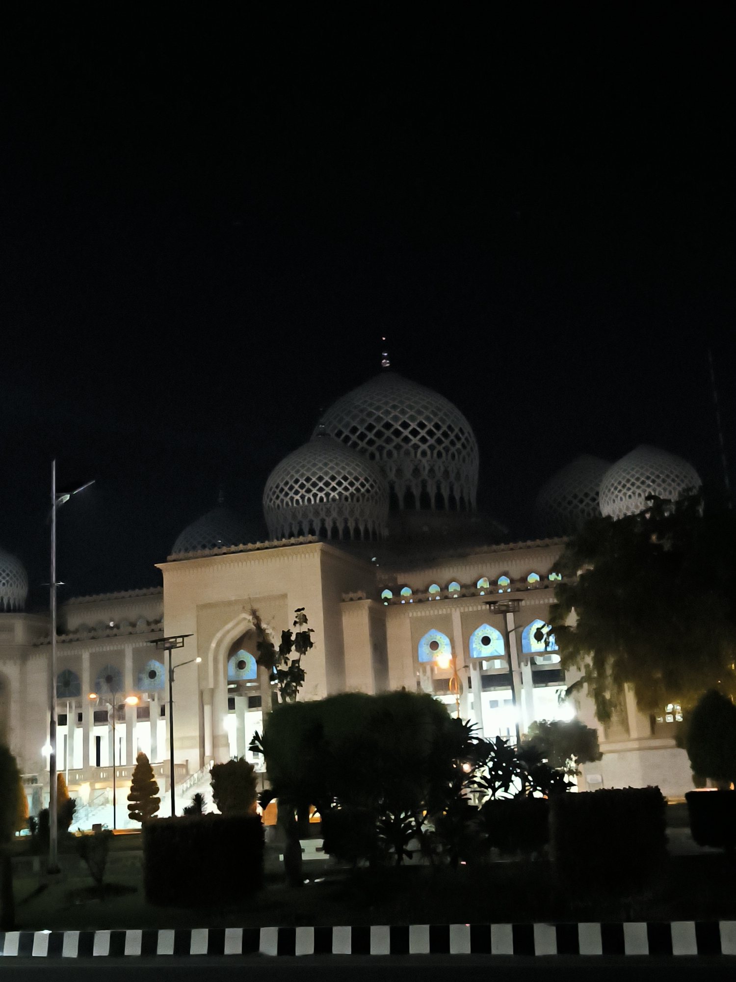 The image depicts the Baiturrahman Grand Mosque in Lhokseumawe, Indonesia, at night. The mosque features multiple domes, with the largest being centrally located and decorated with a lattice pattern. The building is illuminated, highlighting its architectural details. Below the building, there is a manicured landscape with shrubs and trees in the foreground. A black and white striped curb separates the foreground from the mosque's grounds. The sky is dark, suggesting a night time setting.