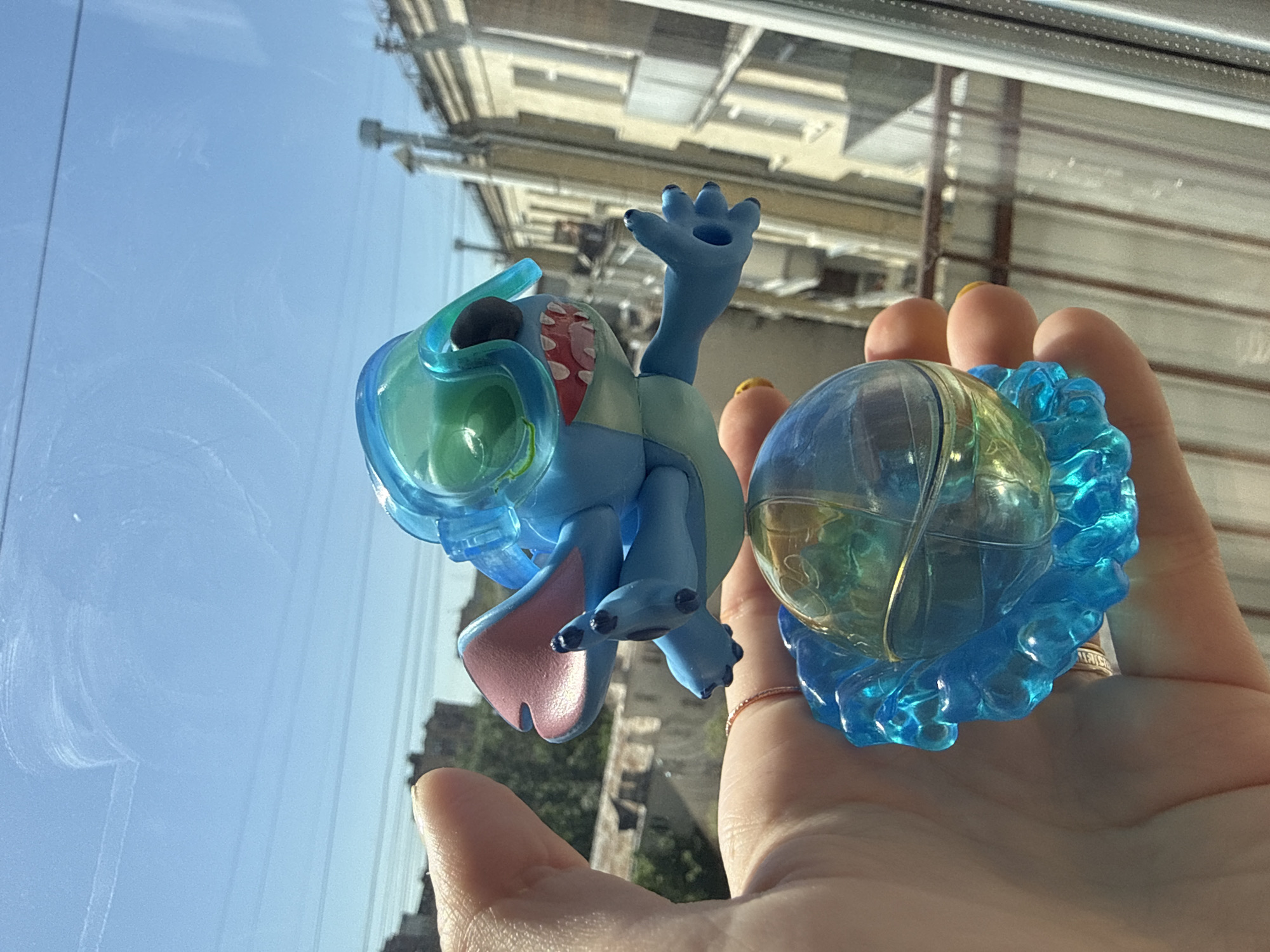 A hand holds a Stitch figurine and a water-filled sphere near a window. Outside the window, a partial view of Moscow's urban landscape is visible.