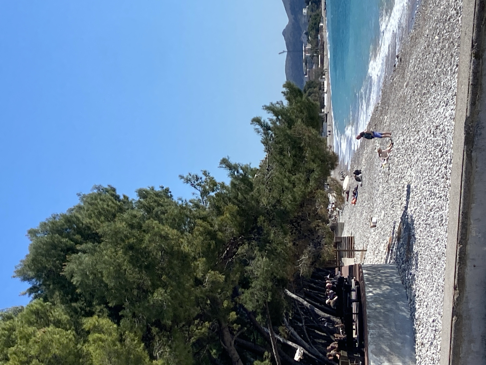 The scene depicts a beach in the city of Bar, Montenegro, composed of smooth, light-colored pebbles. The turquoise sea meets the shore, fringed with white waves. In the upper portion of the image, buildings are visible against the backdrop of a mountain. On the beach, several individuals are present, with one person standing near the water's edge. Another person is lying on the pebbles. A dense cluster of green trees line the beach, partially obscuring a shaded area with wooden benches or structures. The sky is clear and blue. In the foreground, a concrete wall or ledge is visible.