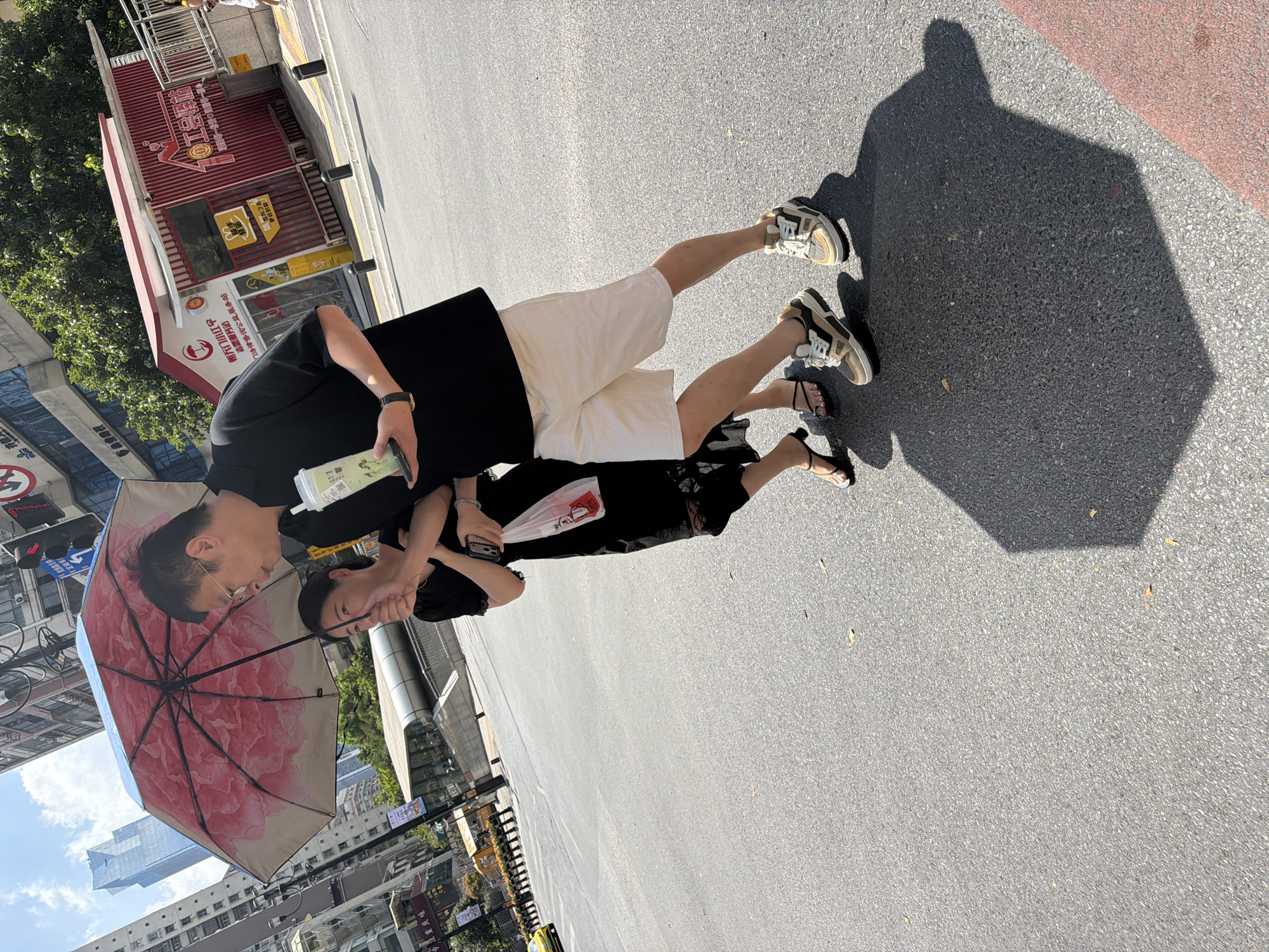 In Yuzhong District, China, a man and a woman walk side-by-side on a paved city street under a large, pink floral umbrella, shielding themselves from the sun. The man, wearing a black polo shirt and light-colored shorts, holds a green beverage cup in his right hand and the umbrella with his left. The woman, dressed in a black lace-trimmed dress, walks closely to his left, holding a white plastic bag with red text. They are walking towards the right of the frame, casting a long shadow on the asphalt. In the background, modern buildings with glass facades rise into a clear blue sky. To the right, a red and white kiosk-like structure with signage is visible, along with trees and streetlights. A yellow vehicle is seen in the distance on the left.