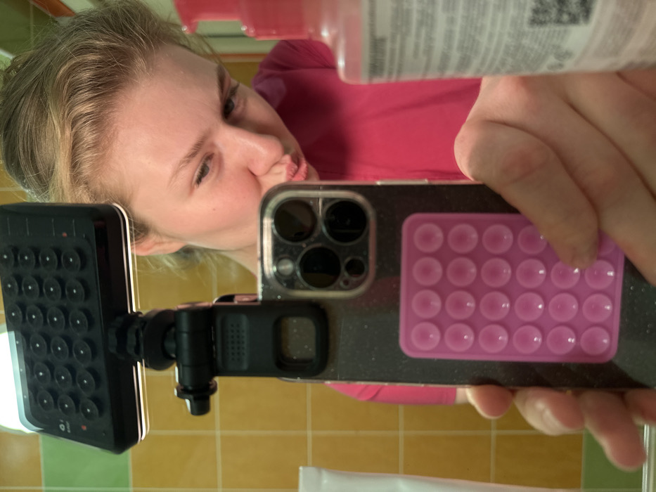A woman with blonde hair is seen in a mirror selfie. She is wearing a pink shirt. Her phone has a dark glittery case with a pink pop-it grip attached. The phone is equipped with a black suction cup mount and a small black video light. The reflection reveals a yellow-tiled wall, suggesting a bathroom setting. The woman is puckering her lips for the photo. A white object is also visible in the reflection near the bottom of the frame.