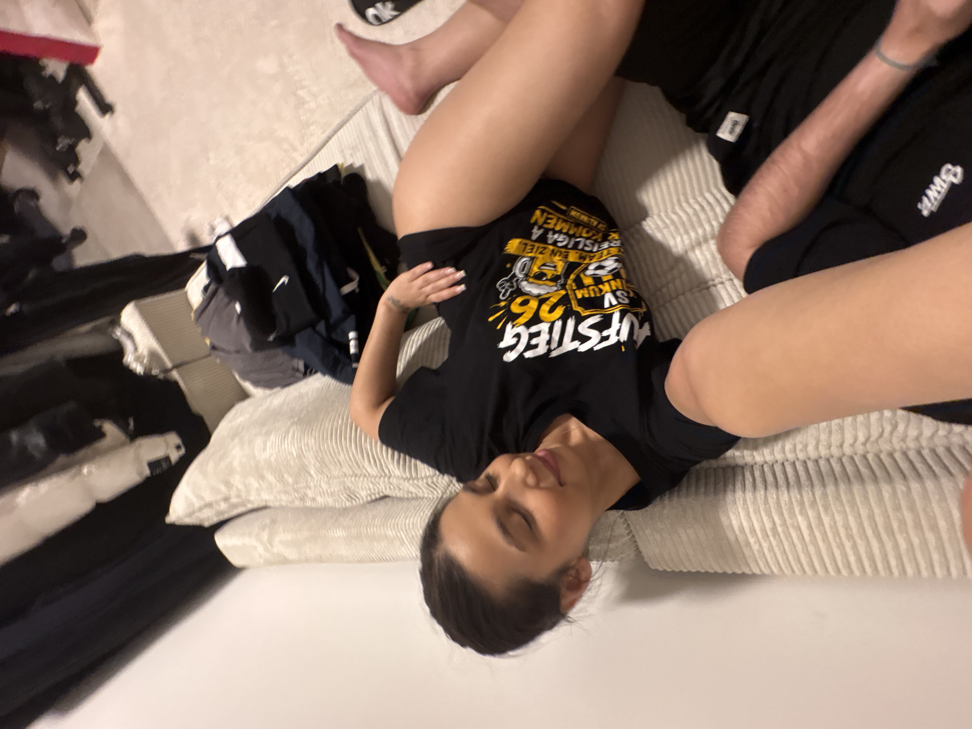 A young woman is lying on a textured, off-white sofa, her body angled towards the camera. She is wearing a black t-shirt with a yellow and black graphic print, the text on which is partially visible. Her left hand rests on her hip. Her legs are exposed from mid-thigh downwards, and her right foot is visible. Next to her, on the sofa, are several folded dark-colored clothing items, some appearing to be t-shirts and shorts, with a black Nike logo visible on one. To the right of the woman, a portion of another person's arm and leg, clad in black shorts, is visible on the sofa. The environment suggests an indoor setting, possibly a living room, with a cream-colored carpet visible in the background and what appears to be clothing hanging on a rack or in a closet in the upper left portion of the image. The overall lighting is dim. The visible text on the woman's t-shirt, "AUFSTIEG 26", and the mention of "REISLIGA A", could potentially link the image to a sporting context in Mönchengladbach, Germany, perhaps related to a football club's promotion.