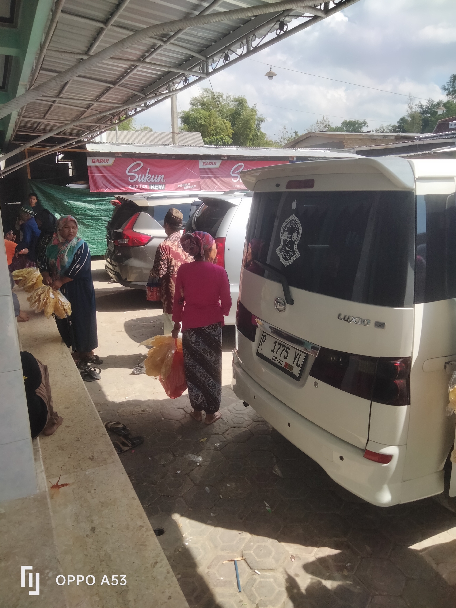 In a street scene in Badung, Indonesia, two white vans are parked next to each other under an open-sided shelter with a corrugated metal roof. The van on the right, a Daihatsu Luxio, has a sticker on its rear window. Several people, including two women in headscarves holding plastic bags, are standing on a paved area next to the vans. A red banner advertising "Sukun KING SIZE NEW" is visible on a wall behind the vans. The ground is paved with hexagonal tiles, and there is some debris scattered on it. The sky is visible in the background with some clouds.