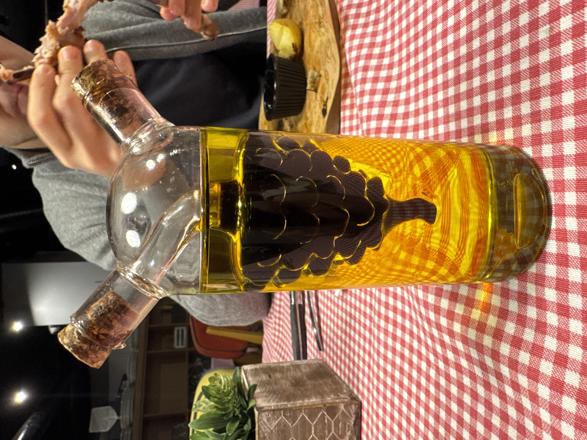 A glass bottle filled with yellow liquid and a dark grape cluster shape inside lies horizontally on a red and white checkered tablecloth. The bottle has two necks with corks. A person is holding what appears to be a bone in their hands. The location cannot be determined from the image.