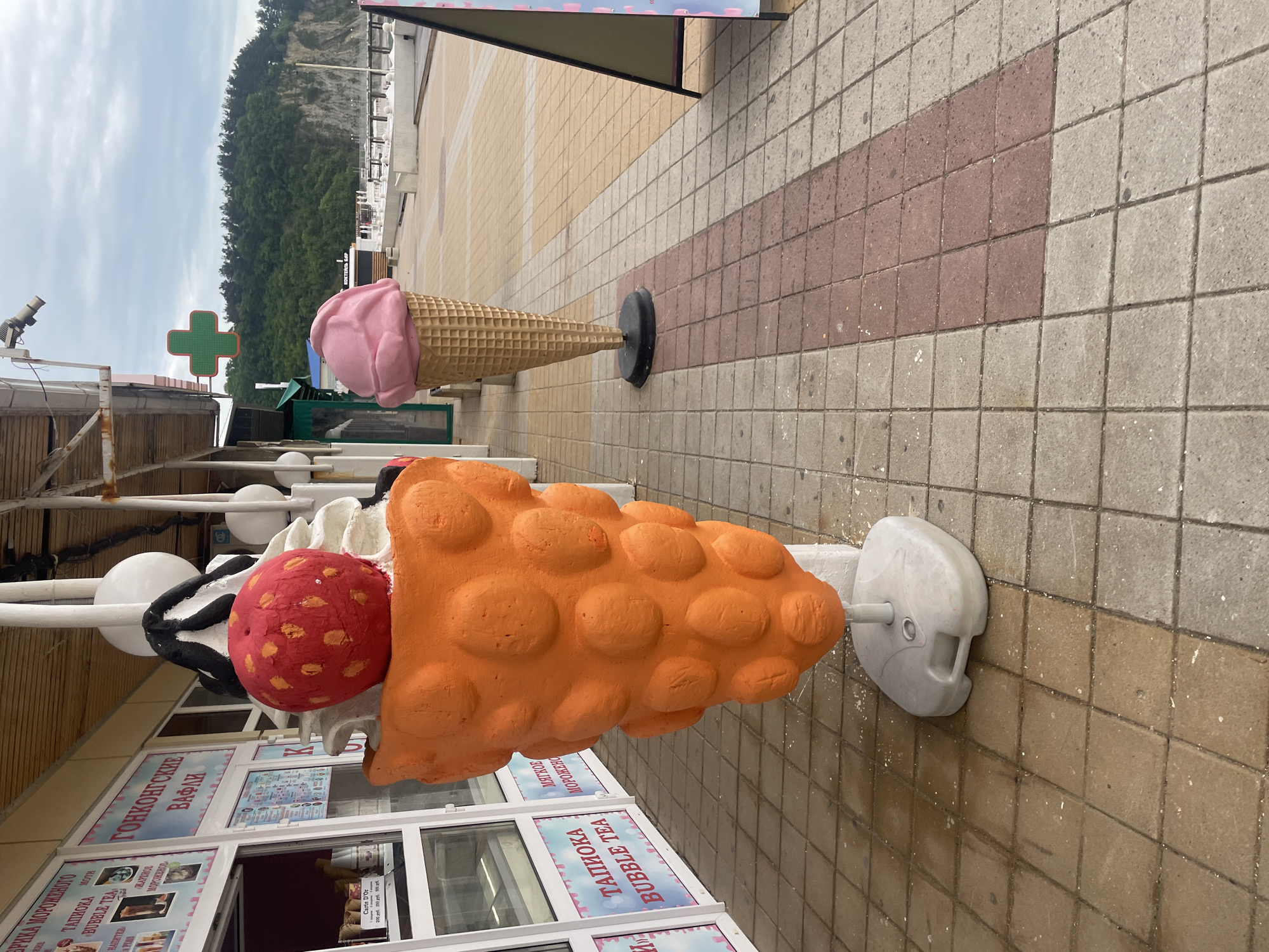 The image shows two oversized, artificial ice cream cones displayed outside a building with Russian language signage, likely a dessert or ice cream shop. The shop’s name includes the phrases “Bubble Tea” and “Hong Kong Waffles.” The foreground cone is orange and topped with a red sphere resembling a fruit. The background cone is pink and more traditionally shaped. The location is likely in Olginka, Russia. The setting appears to be a pedestrian walkway or street with tiled surfaces. In the distance, a treelined hillside or mountain is visible. A green cross, likely indicating a pharmacy, is also visible.