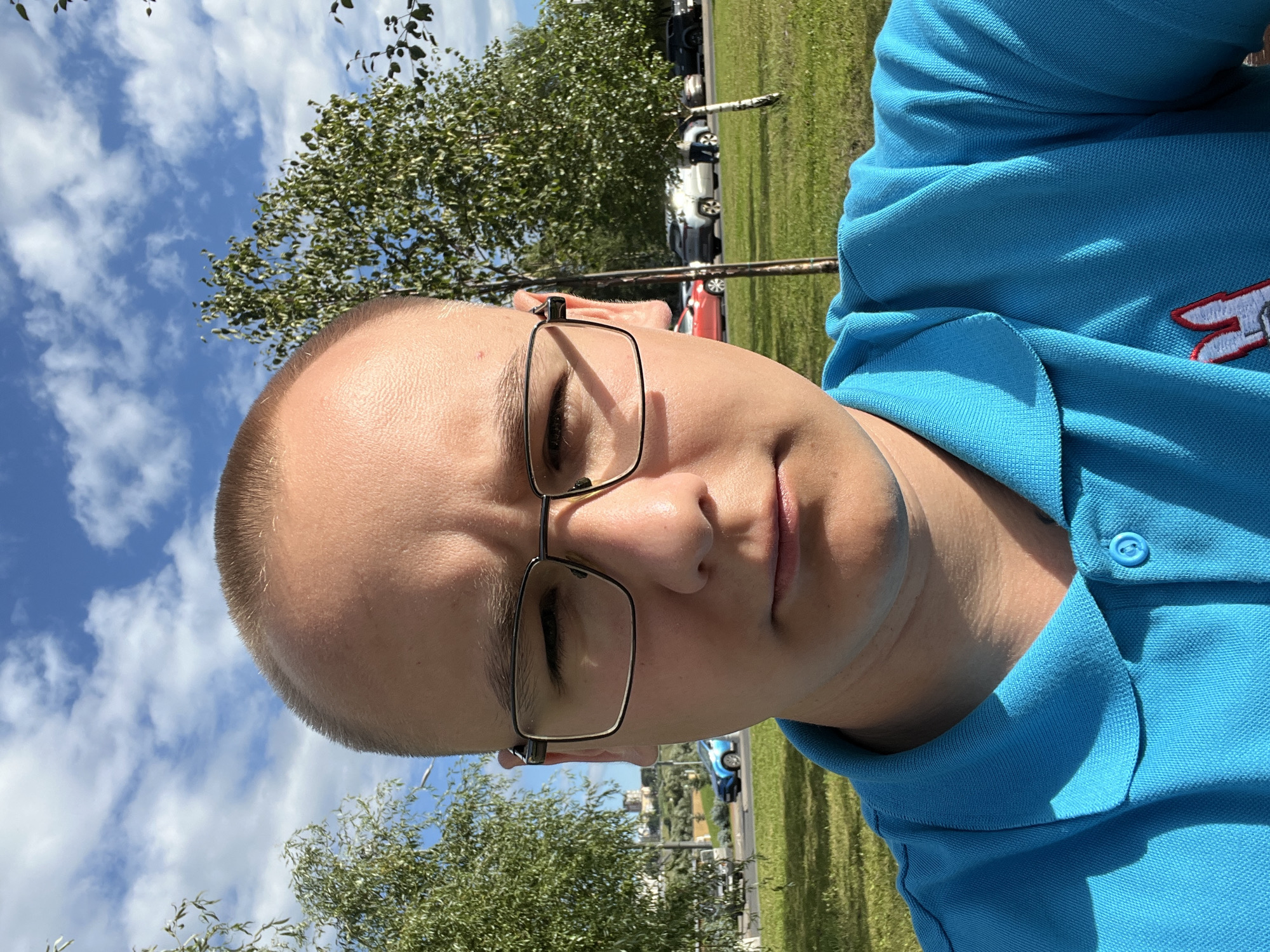 A young man with a buzz cut and glasses is taking a selfie outdoors. He is wearing a bright blue polo shirt with a small embroidered logo on the left chest. He is looking directly at the camera with a neutral expression. The background shows a park-like setting with green grass, trees, and a bright blue sky with scattered white clouds. In the distance, there is a road with parked cars, including a blue vehicle. This scene is likely located in a public park within the city of Nizhny Novgorod, Russia.