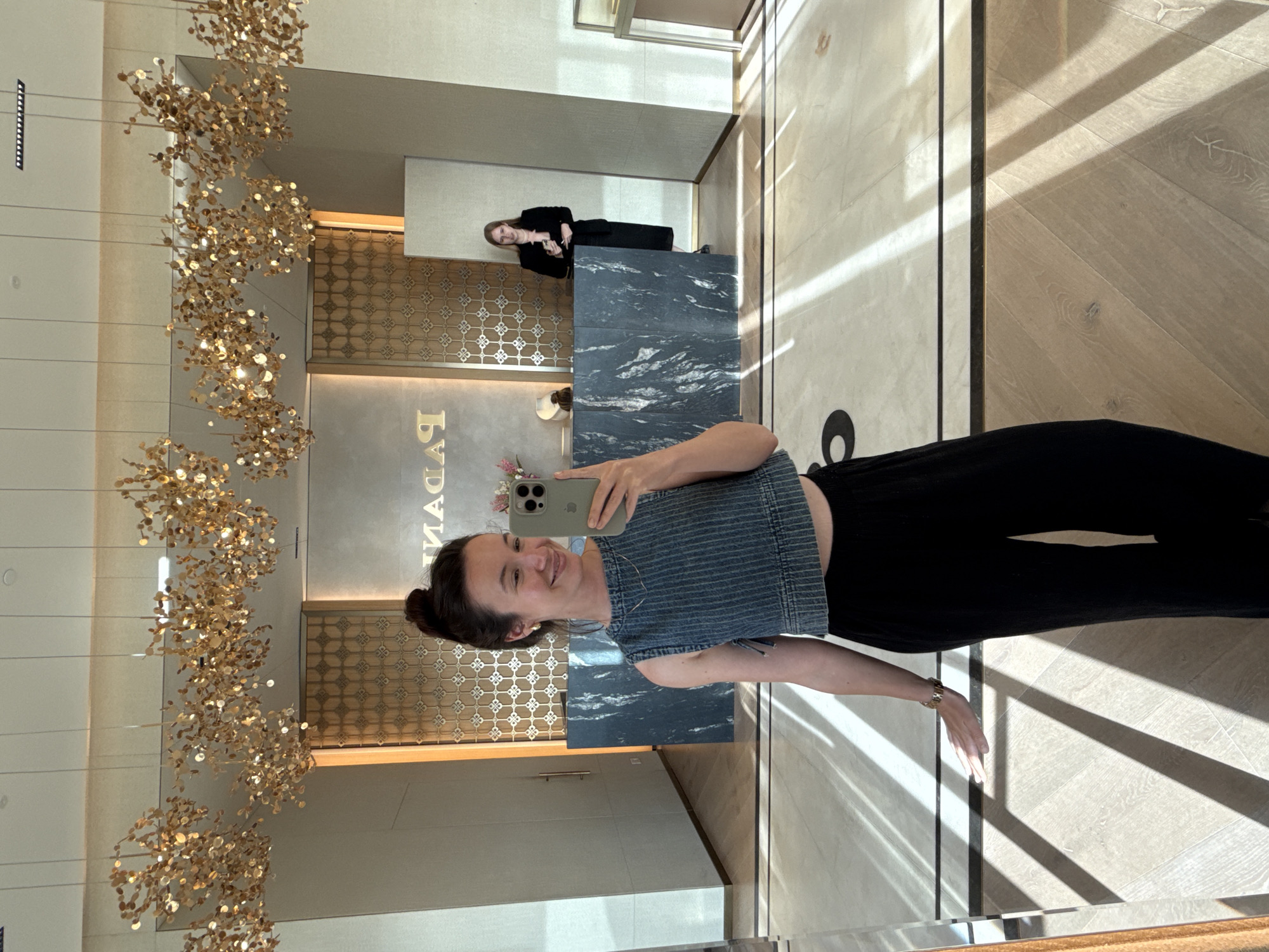 The image shows a woman taking a selfie in a high-end jewelry store. She is wearing a denim crop top and black pants. In the background, there is a reception desk with a dark, marbled surface and a gold-patterned wall with the store's name, "PAGANI," visible. Another woman, likely an employee, stands behind the reception desk. The store features modern chandeliers with gold accents and a polished marble floor. The sunlight creates strong diagonal shadows across the floor. The environment suggests a luxurious retail space.
