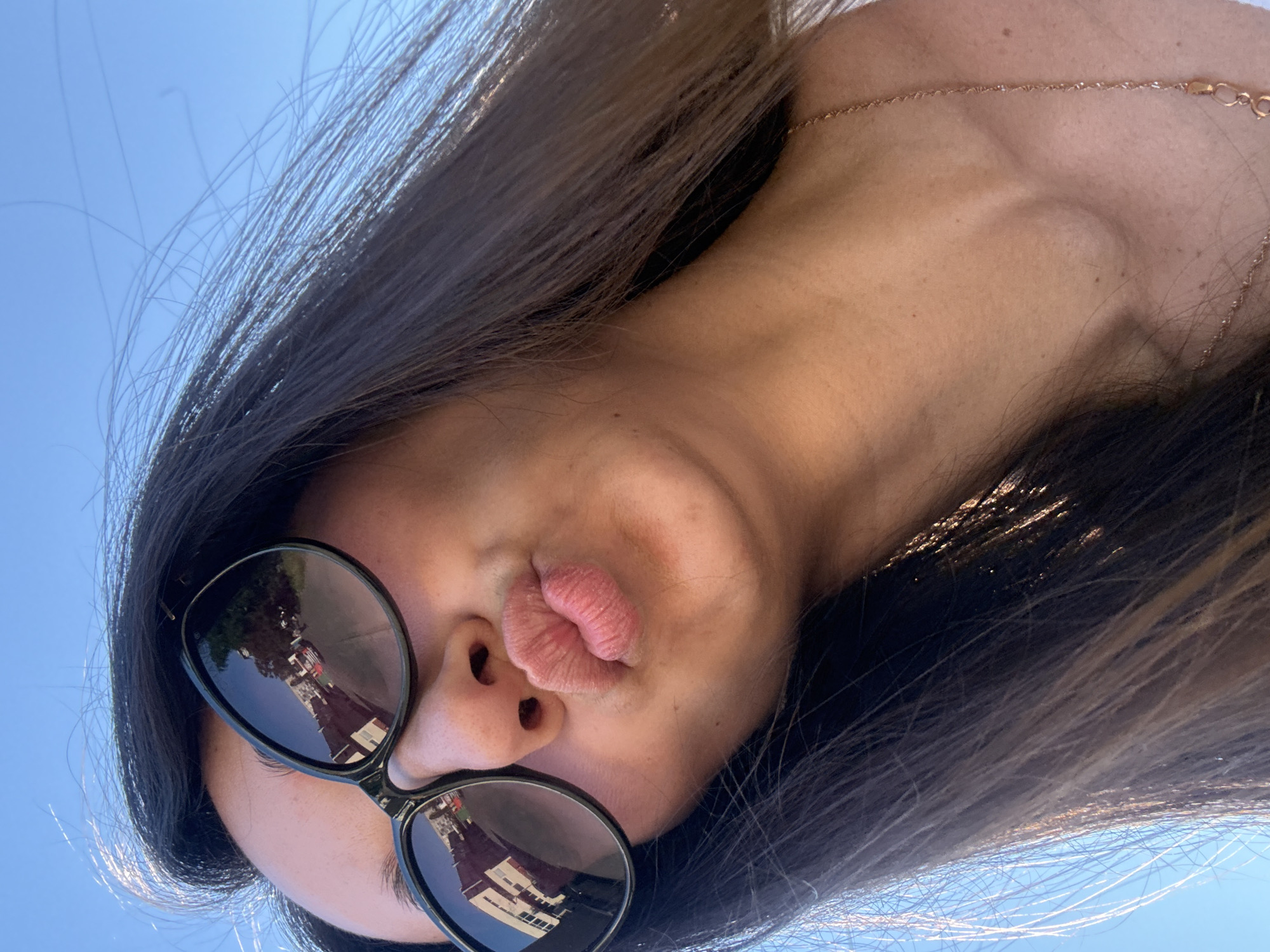 A close-up, low-angle, outdoor selfie shows a woman with dark hair, wearing black sunglasses and a necklace, making a kissing face. The background is a clear blue sky. The sunglasses reflect buildings and trees, suggesting an urban environment. The image is a selfie taken from a low angle, looking up at the subject. The woman's hair is blowing in the wind. Her lips are pursed in a kiss. A thin gold chain necklace is visible around her neck. The lighting suggests a sunny day. No specific location in Kyiv, Ukraine is identifiable from the image.
