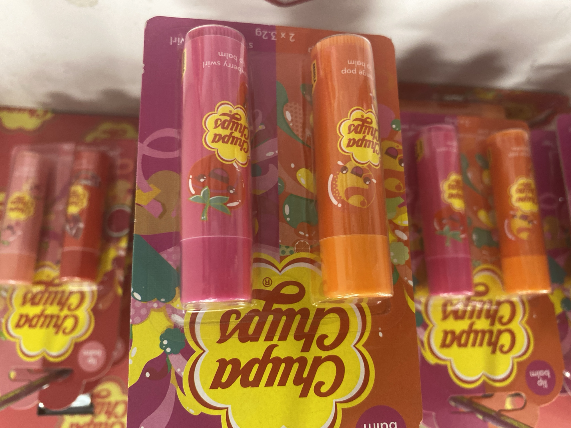 The image shows a retail display of Chupa Chups lip balms. The lip balms are packaged in pink and orange tubes with cartoonish fruit designs and the Chupa Chups logo. The display is set against a white background, suggesting it is likely in a store. The lighting is bright, indicating an indoor setting. There are no people visible in the image. The overall impression is of a colorful and playful product advertisement. The location is not identifiable as Konya, Turkey, or any other specific city.