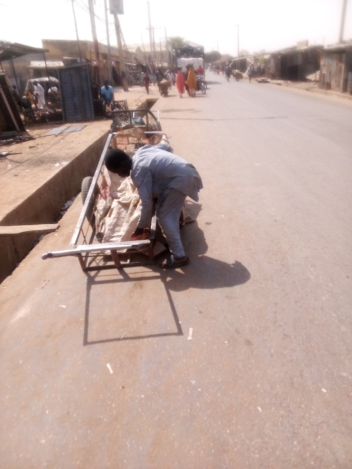 The image depicts a daytime street scene in Tudun Murtala, Nigeria, under bright sunlight with atmospheric haze. In the foreground, a man with dark hair, wearing a light grey long-sleeved tunic and trousers with dark sandals, is bent over a metal-framed two-wheeled pushcart. He is positioned with his back partially to the viewer, facing right, with his hands near the cart's contents. The cart contains multiple light-colored sacks, appearing to be made of fabric or burlap. The cart is situated on a concrete surface next to a concrete drainage channel on the left, with its right side on a light brown paved road.
The paved road extends into the background, flanked on the left by various structures including market stalls and buildings constructed from corrugated metal and masonry. Several individuals are visible in the midground: one person in a blue garment near the left-side structures, and two individuals in red and yellow/cream garments walking away from the viewer on the left side of the road. Another person is pushing a two-wheeled cart further down the street. A large truck, covered with a dark tarp, is visible further down the street on the left. Utility poles with wires line the left side of the street. The background shows the road receding, bordered by similar structures, under a bright, pale sky.