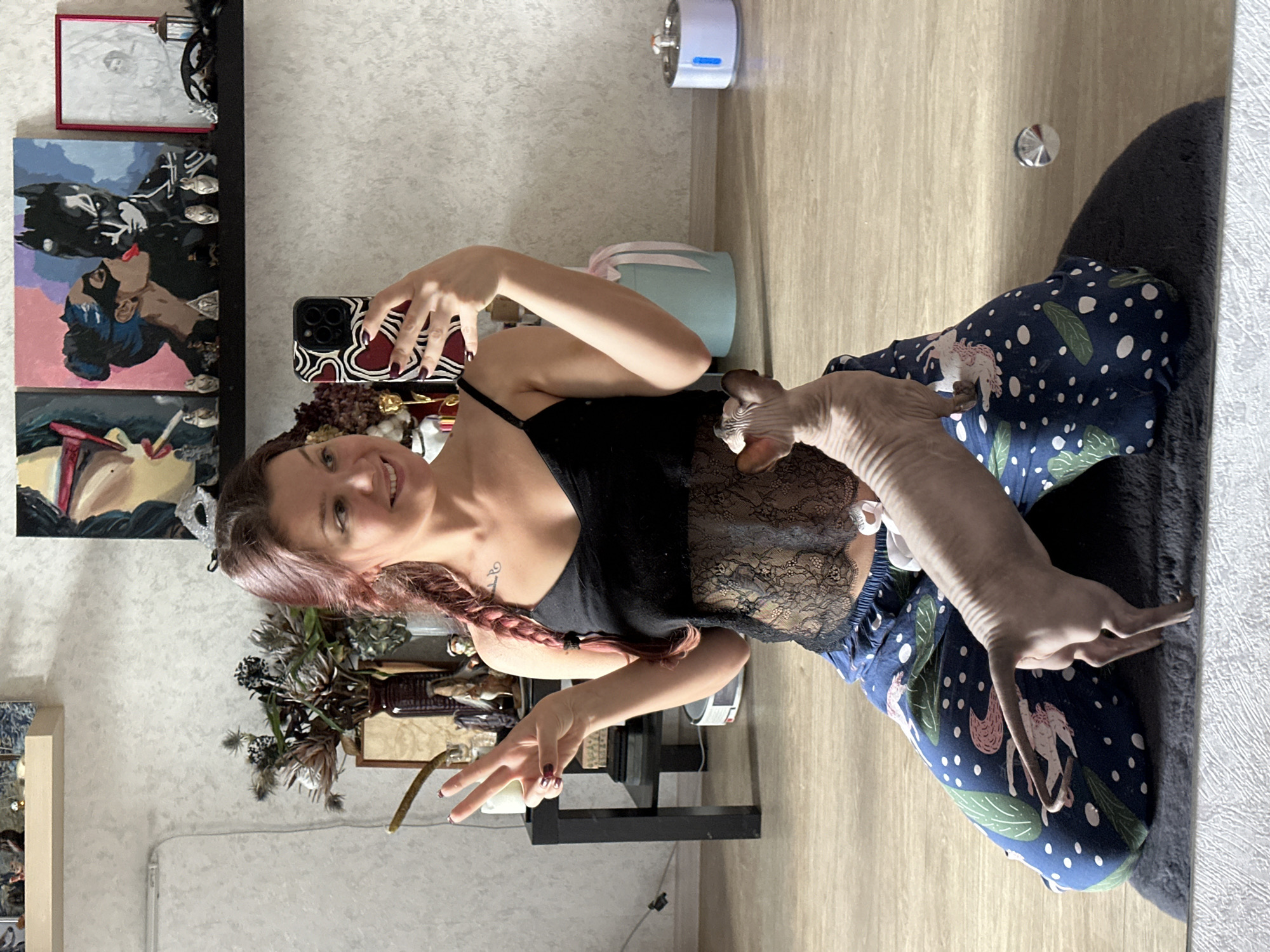 The scene is an indoor shot, likely a selfie, featuring a woman seated on a dark blue rug against a light wooden floor. The woman is fair-skinned with reddish-brown hair styled in a braid. She is wearing a black top and patterned blue pants. She holds a smartphone in her right hand, possibly taking the picture, and displays a "peace" sign with her left hand. Beside her sits a Sphynx cat on her lap.
The background is a mix of domestic elements. On the left, artwork is displayed on the wall along with floral arrangements and decorations on shelves. A cylindrical object is visible on the top right of the frame, mounted to the wall.
There is no direct evidence to suggest the location is Vladimir, Russia, from the image itself. Additional contextual information would be needed to confirm such a location.